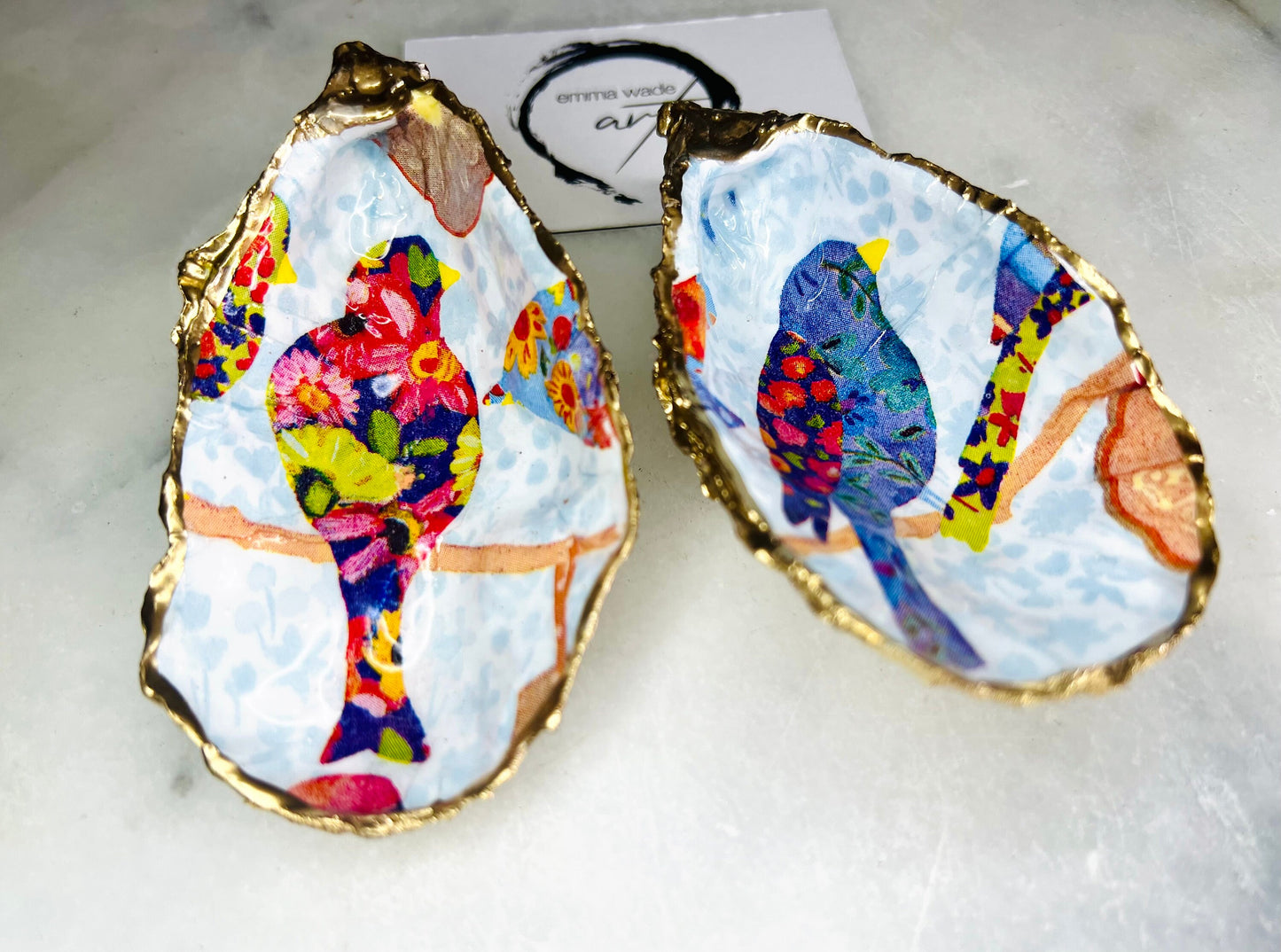 Handmade Dorset oyster shell trinket dishes featuring colourful bird artwork in pink and blue tones, finished with hand-painted gold leaf edging and glossy resin. Decorative coastal jewellery dishes styled on a white marble background with vibrant nature-inspired bird illustrations.