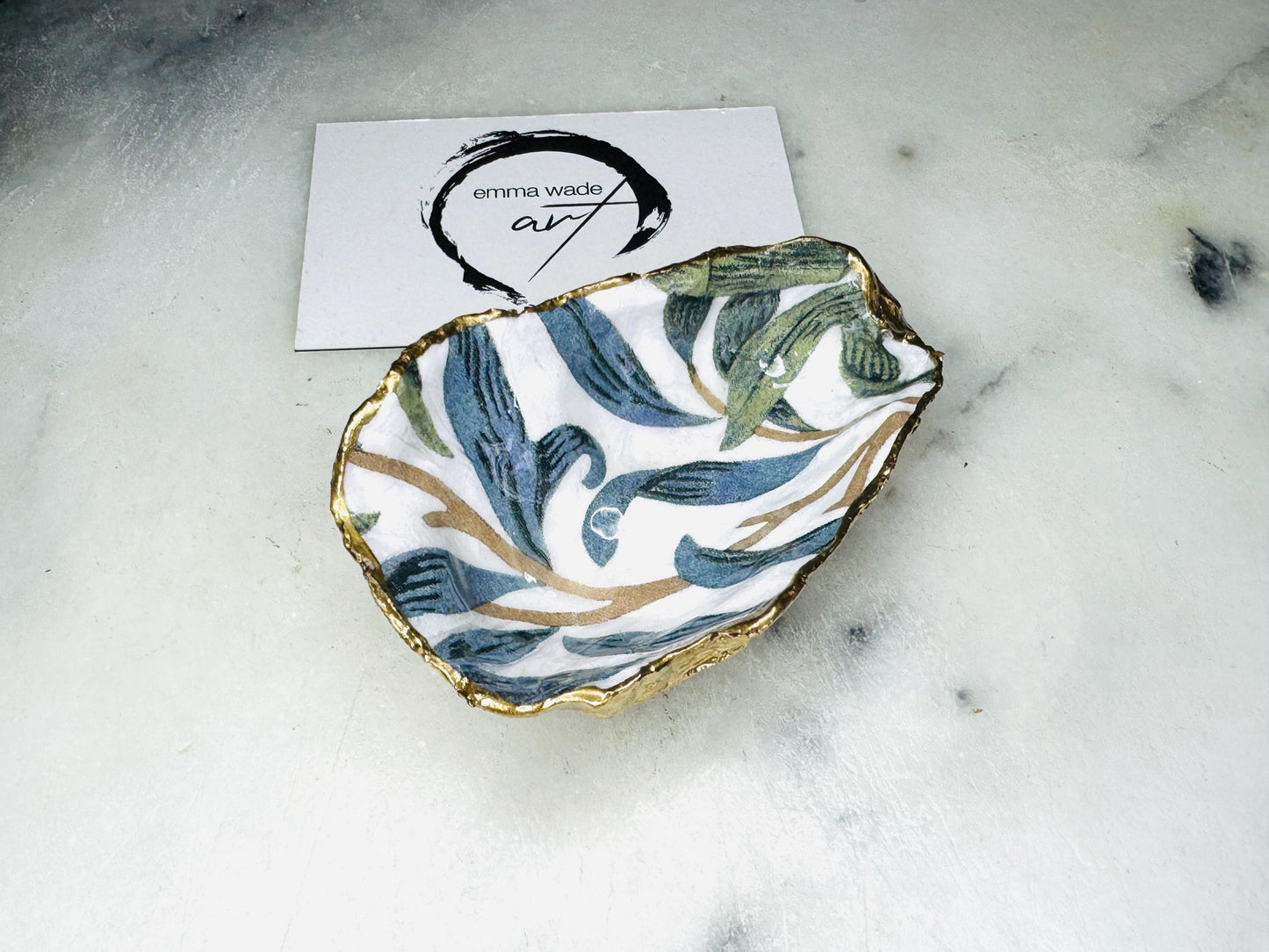 Handmade Dorset oyster shell trinket dish featuring William Morris Willow Bough botanical design in soft blue and sage green tones. Decorative jewellery holder finished with hand-applied liquid gold leaf edging and glossy resin. Coastal heritage décor shell dish styled as a ring holder and elegant bathroom accessory with natural oyster shell shape.