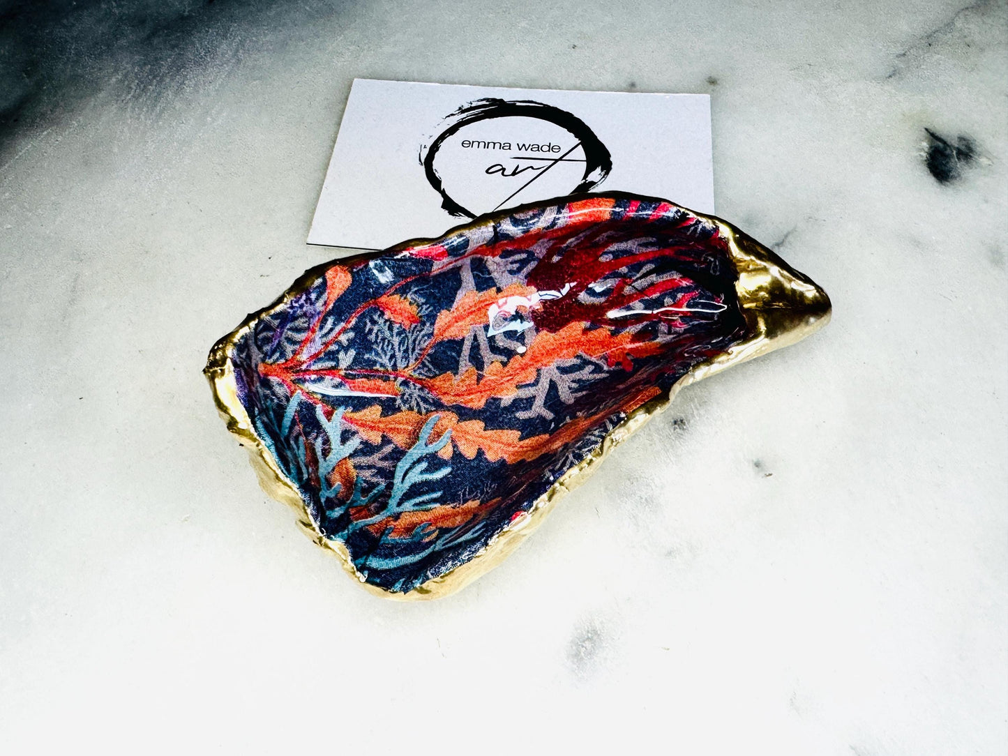 Handmade Dorset oyster shell trinket dishes featuring vibrant sea coral inspired artwork in coral red, navy and turquoise tones, finished with hand-painted gold leaf edging and glossy crystal clear resin. Coastal jewellery dishes styled on a white marble background with rich ocean-inspired botanical patterns and luxurious gilded edges.