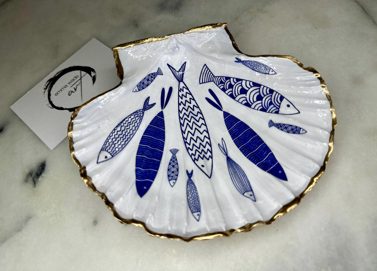 Handmade Dorset scallop shell trinket dishes featuring elegant blue fish illustrations with hand-painted gold leaf edging and glossy crystal clear resin. Coastal nautical jewellery dishes inspired by Scandinavian ocean decor, styled on a neutral background with detailed blue fish artwork and luxurious gilded edges.