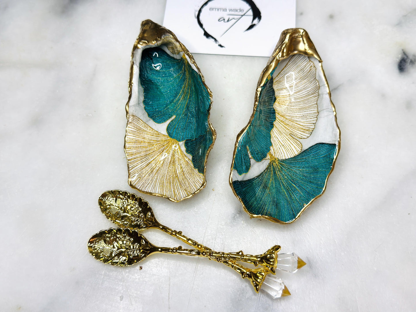A pair of green and gold Japanese Ginkgo oyster shell salt and pepper pinch pots with floral botanical leaf design, hand gilded with liquid gold leaf and sealed in glossy resin. Coastal Dorset oyster shell dishes styled with matching gold crystal spoons on a marble background. Handmade luxury coastal dining décor and wedding gift.