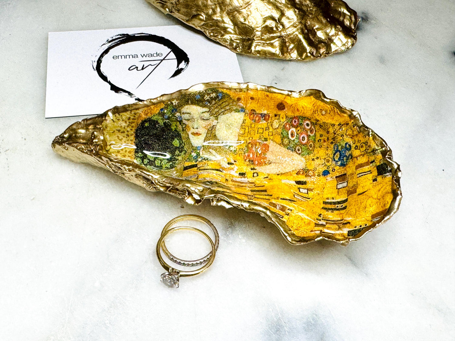 Handcrafted Dorset oyster shell trinket dish inspired by Gustav Klimt’s The Kiss, featuring romantic gold and green artwork detail. Natural oyster shell edged in hand applied liquid gold leaf and sealed with high gloss resin. Luxury art inspired jewellery holder displayed with gold rings on marble background. Unique gift for her or art lover keepsake.