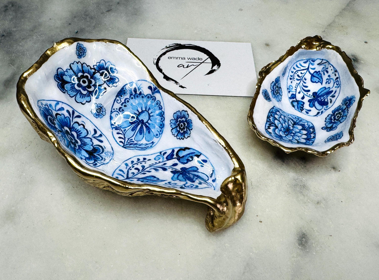 Handmade Dorset oyster shell trinket dish featuring a vintage blue and white decorative egg design with intricate floral porcelain style detailing. Oyster shell is hand gilded with liquid gold leaf edging and sealed in glossy crystal clear resin. Elegant coastal jewelry holder, ring dish, trinket tray, or decorative keepsake inspired by antique ceramics and vintage decor styling. Luxury handmade shell art photographed on a marble background.