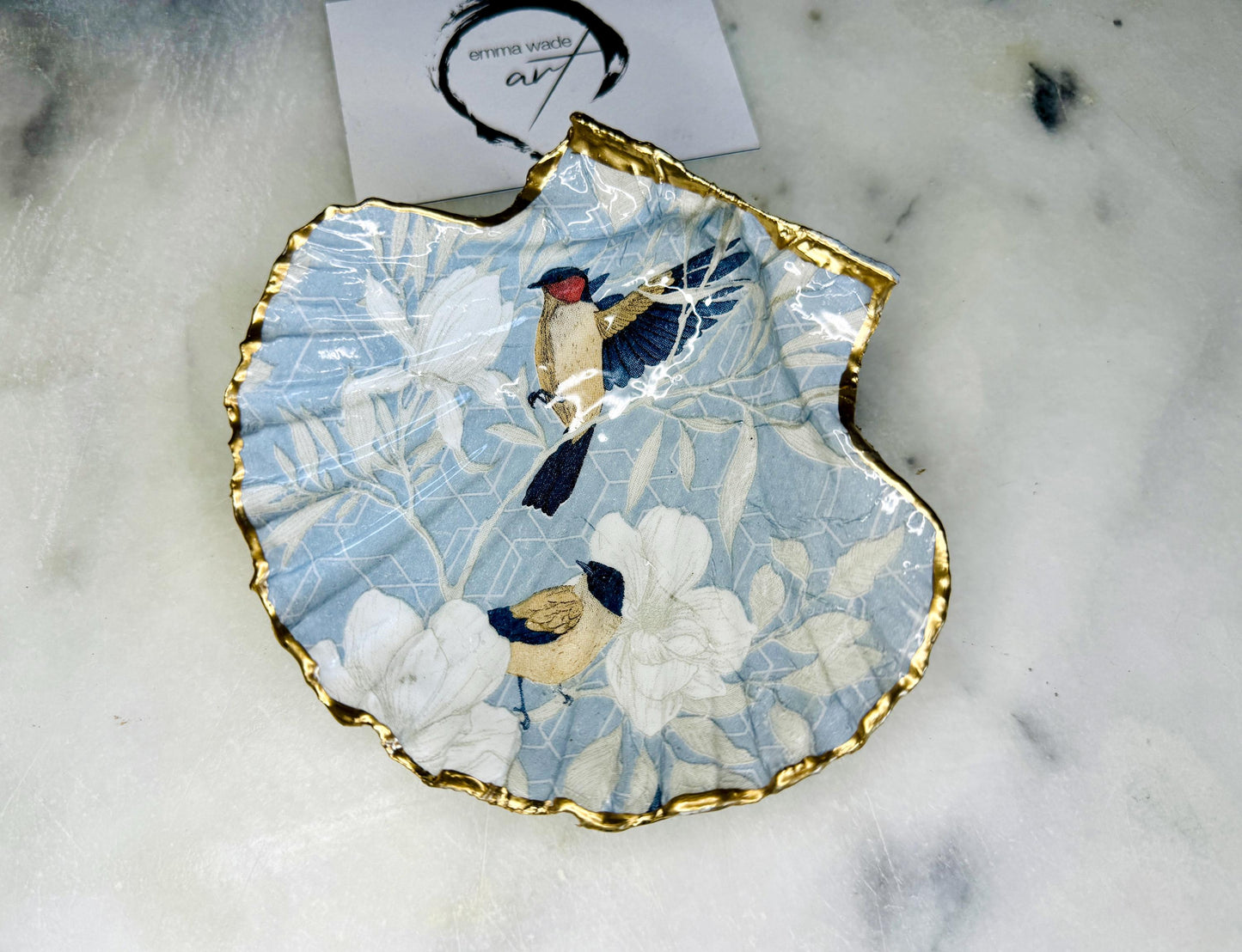 Handmade scallop shell trinket dish featuring a Blue Birds of Paradise decoupage design, finished with real gold leaf and sealed with glossy resin. Crafted from a natural Dorset scallop shell and shown as a jewellery and ring holder on a light background. Coastal-inspired home decor, perfect for holding rings, earrings and small keepsakes, with each shell one of a kind and finished by hand.