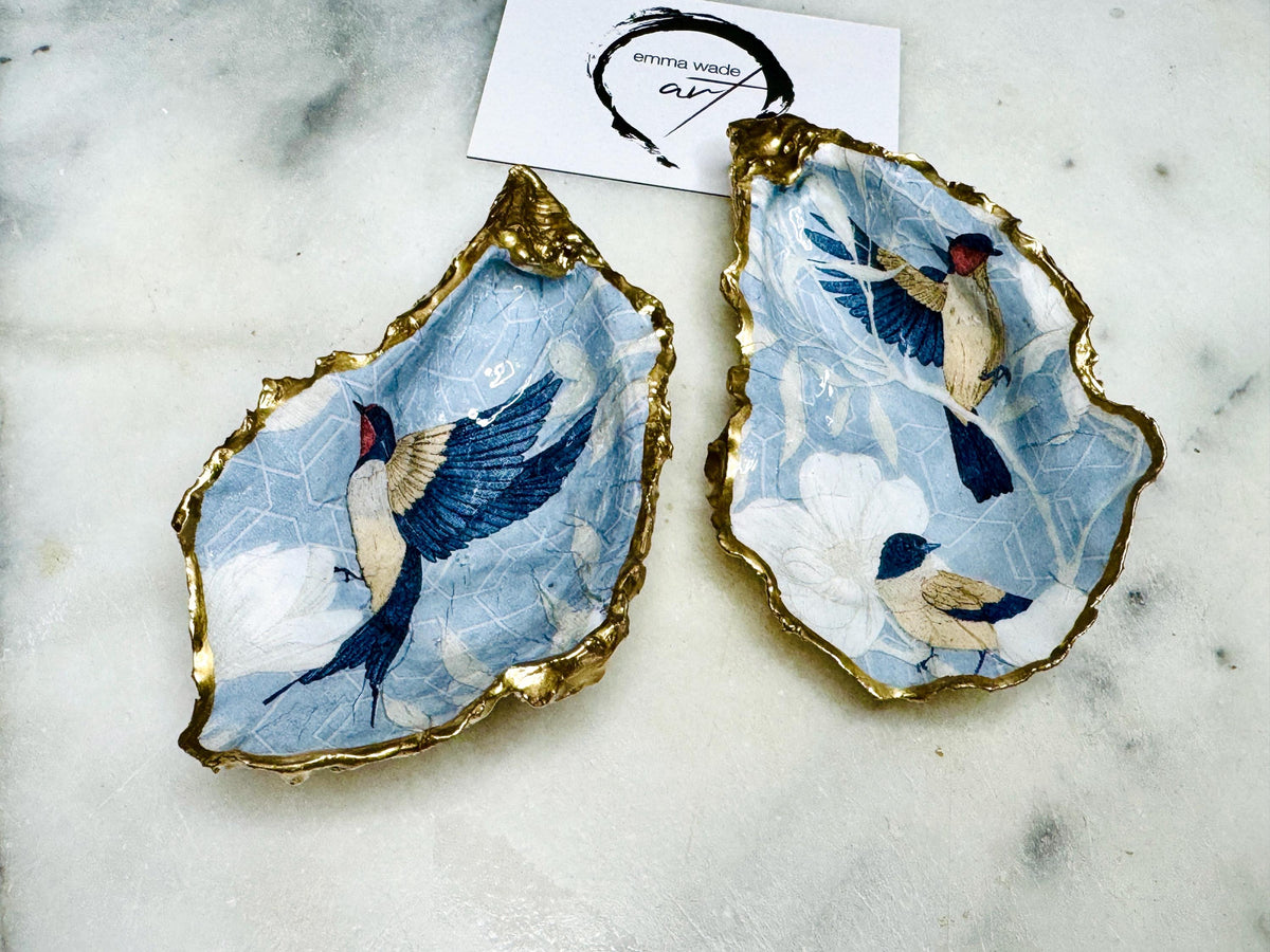 Two natural oyster shell salt and pepper dishes decorated with a blue bird decoupage design, finished with hand-gilded gold leaf edges and sealed with glossy resin. These handmade coastal shell pinch pots are crafted from Dorset shells and styled as an elegant table setting accessory or unique gift.