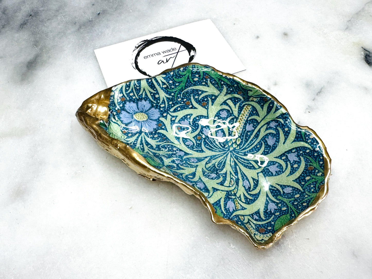 Handmade William Morris inspired oyster shell trinket dish in green and blue tones, finished with gold leaf and sealed with glossy resin. Coastal jewellery dish made from a Dorset oyster shell, featuring a seaweed decoupage design. Perfect for holding rings, earrings and small treasures. Decorative shell bowl photographed on a light background, showing the natural shell shape, detailed pattern and gilded edges.