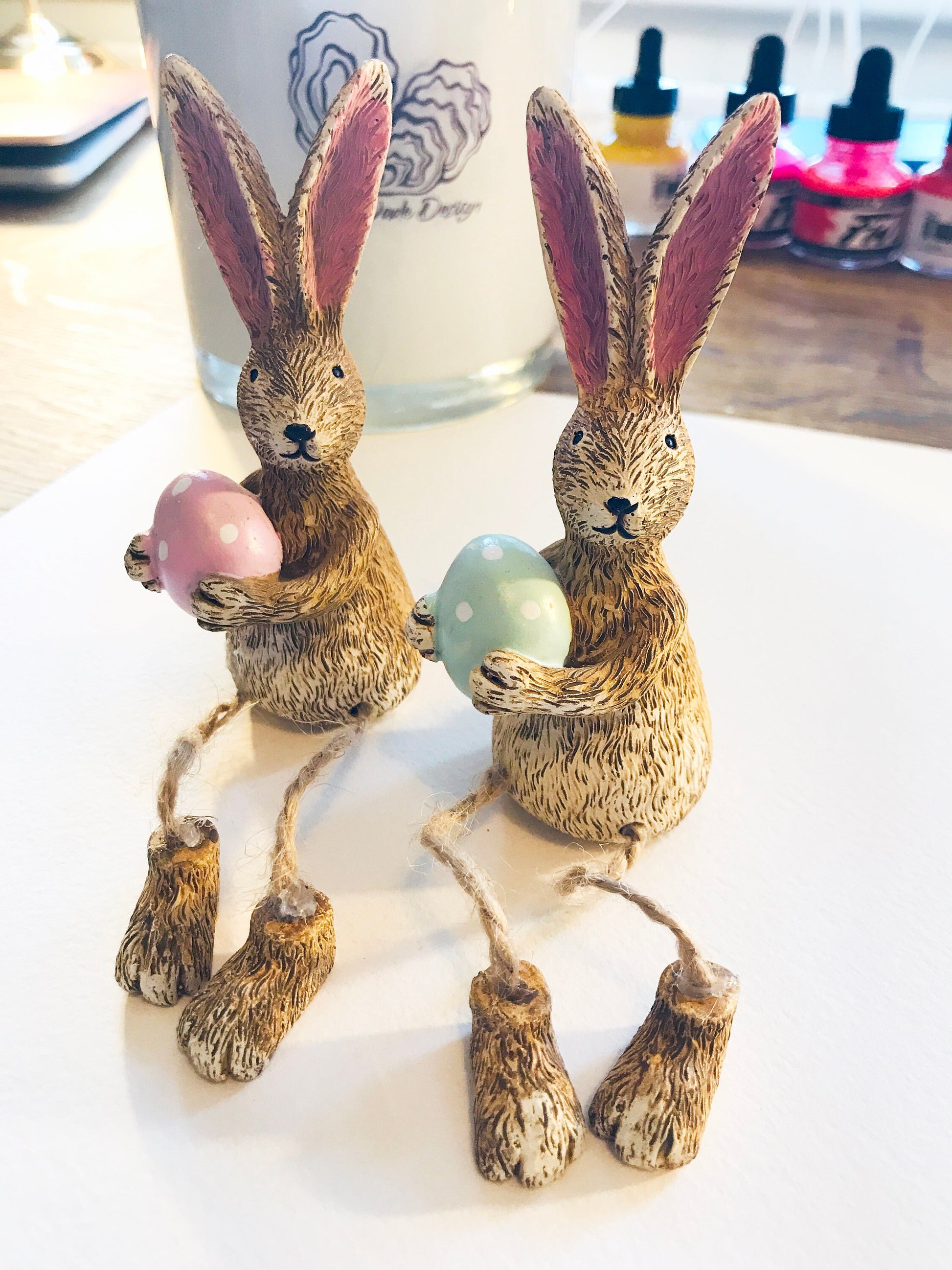 Hand-finished shelf sitting bunny ornament with dangling jute legs and polka dot egg. Available in pink or green. Perfect Easter rabbit shelf decoration, birthday gift, or home décor accessory. Cute animal figurine for rabbit lovers, Easter decorations, new home presents, or seasonal mantel styling.
