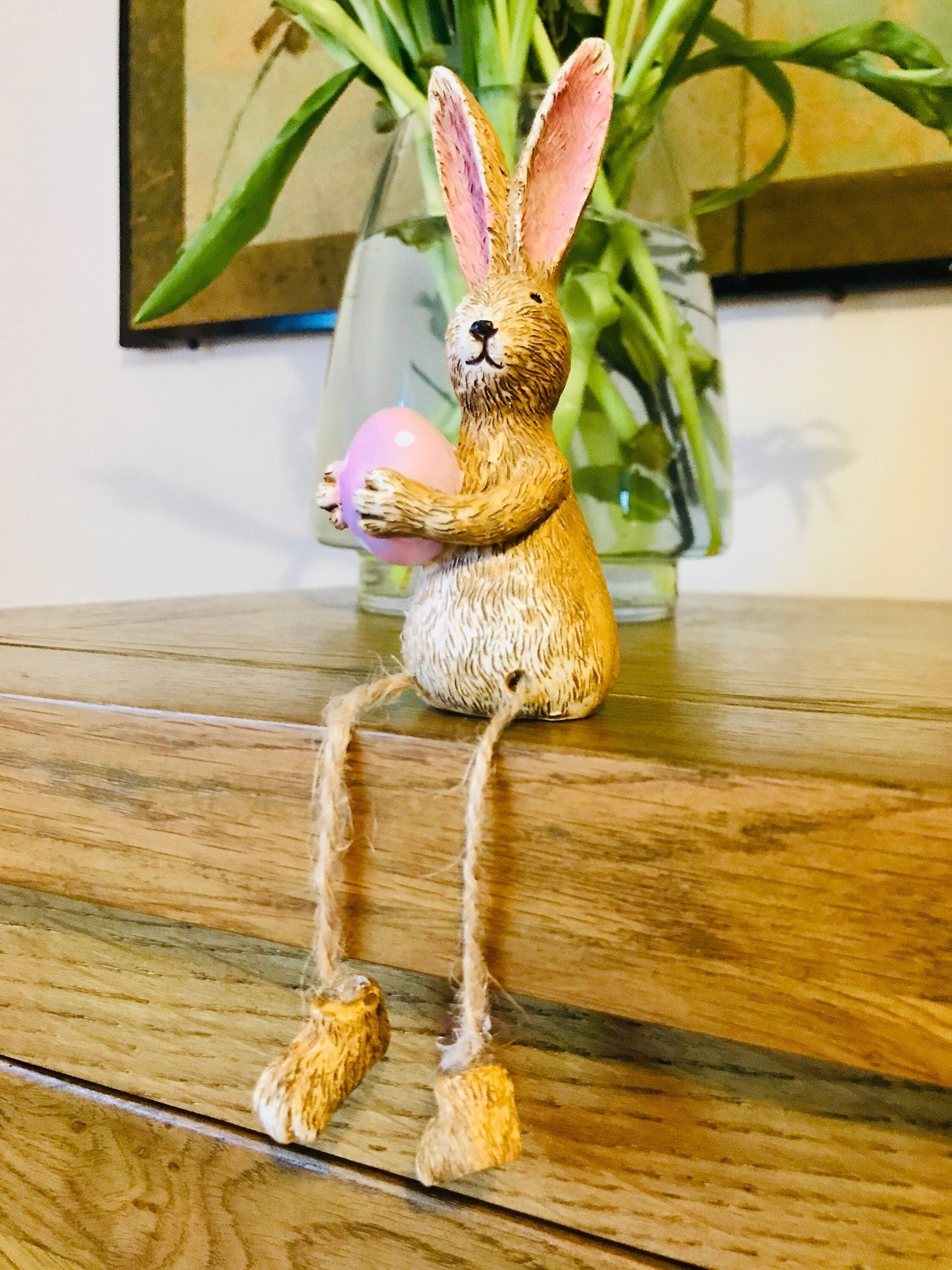 Hand-finished shelf sitting bunny ornament with dangling jute legs and polka dot egg. Available in pink or green. Perfect Easter rabbit shelf decoration, birthday gift, or home décor accessory. Cute animal figurine for rabbit lovers, Easter decorations, new home presents, or seasonal mantel styling.