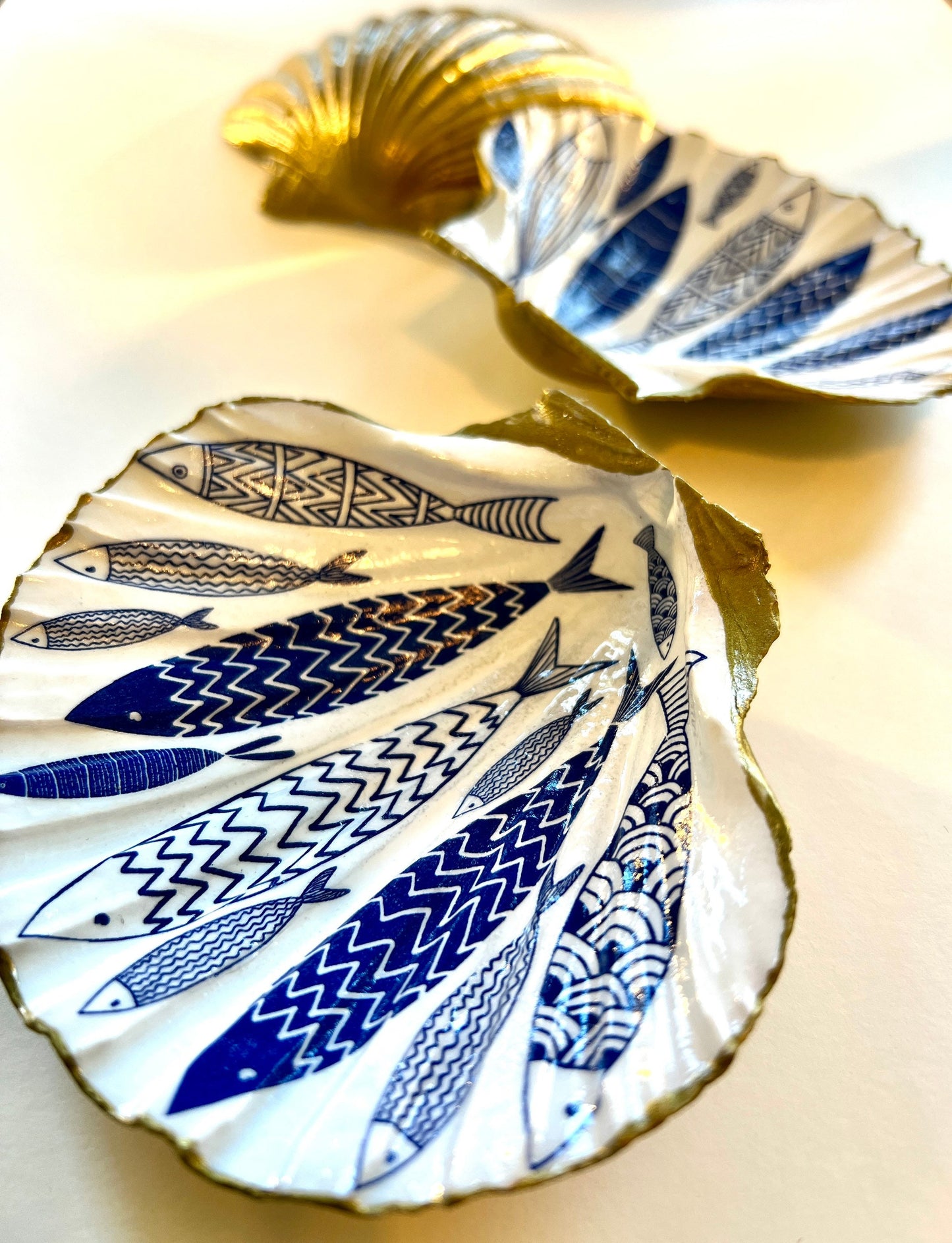 Handmade Dorset scallop shell trinket dishes featuring elegant blue fish illustrations with hand-painted gold leaf edging and glossy crystal clear resin. Coastal nautical jewellery dishes inspired by Scandinavian ocean decor, styled on a neutral background with detailed blue fish artwork and luxurious gilded edges.
