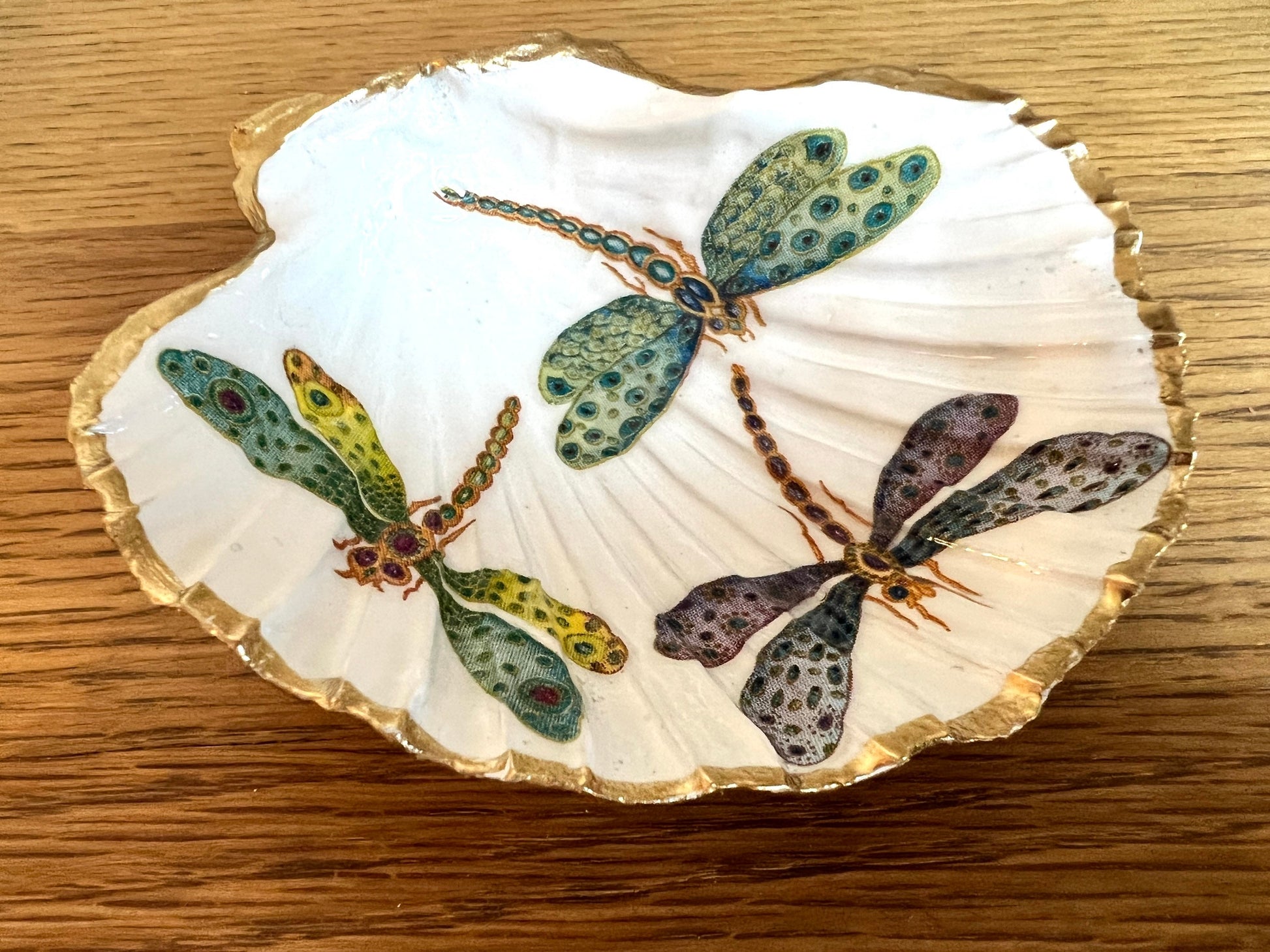 A handmade scallop shell trinket dish decorated with a blue and green dragonfly design and finished with gold leaf, sealed with a glossy clear resin. The natural ridges and curved shape of the shell are visible beneath the painted and decoupaged pattern, giving the piece texture and depth. The dish is shown as a small decorative tray suitable for holding rings, jewellery, or keys and works as a coastal and nature-inspired accent in a bedroom, bathroom, or dressing area.