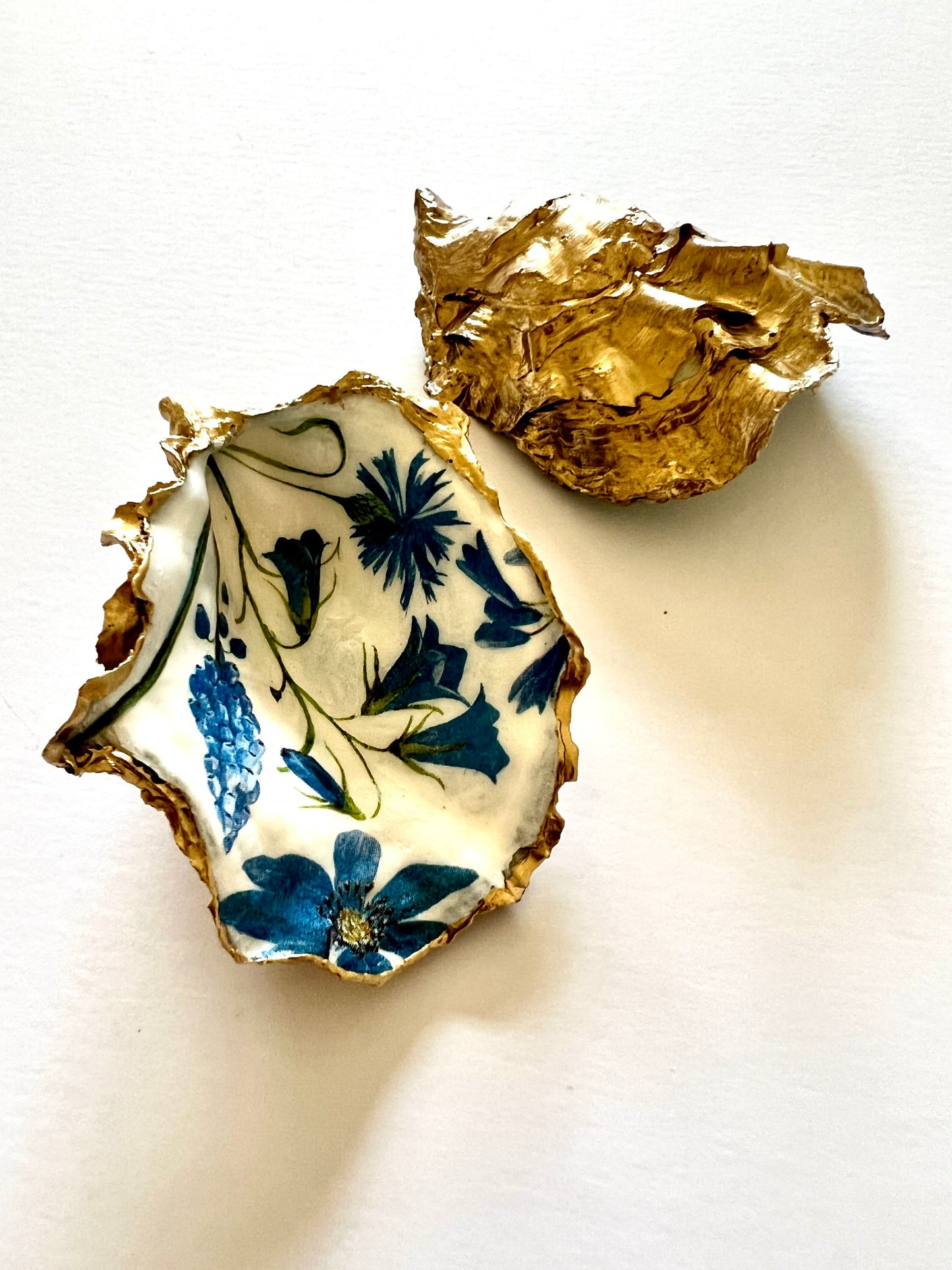 Handmade Dorset oyster shell trinket dish featuring delicate blue floral artwork including bluebells and forget-me-nots, finished with hand-applied gold leaf edging and glossy resin. Coastal-inspired jewellery dish and ring holder available in multiple sizes including XL oyster shells. Elegant handmade home décor gift ideal for birthdays, bridesmaids, Mother’s Day, weddings, and coastal interiors.