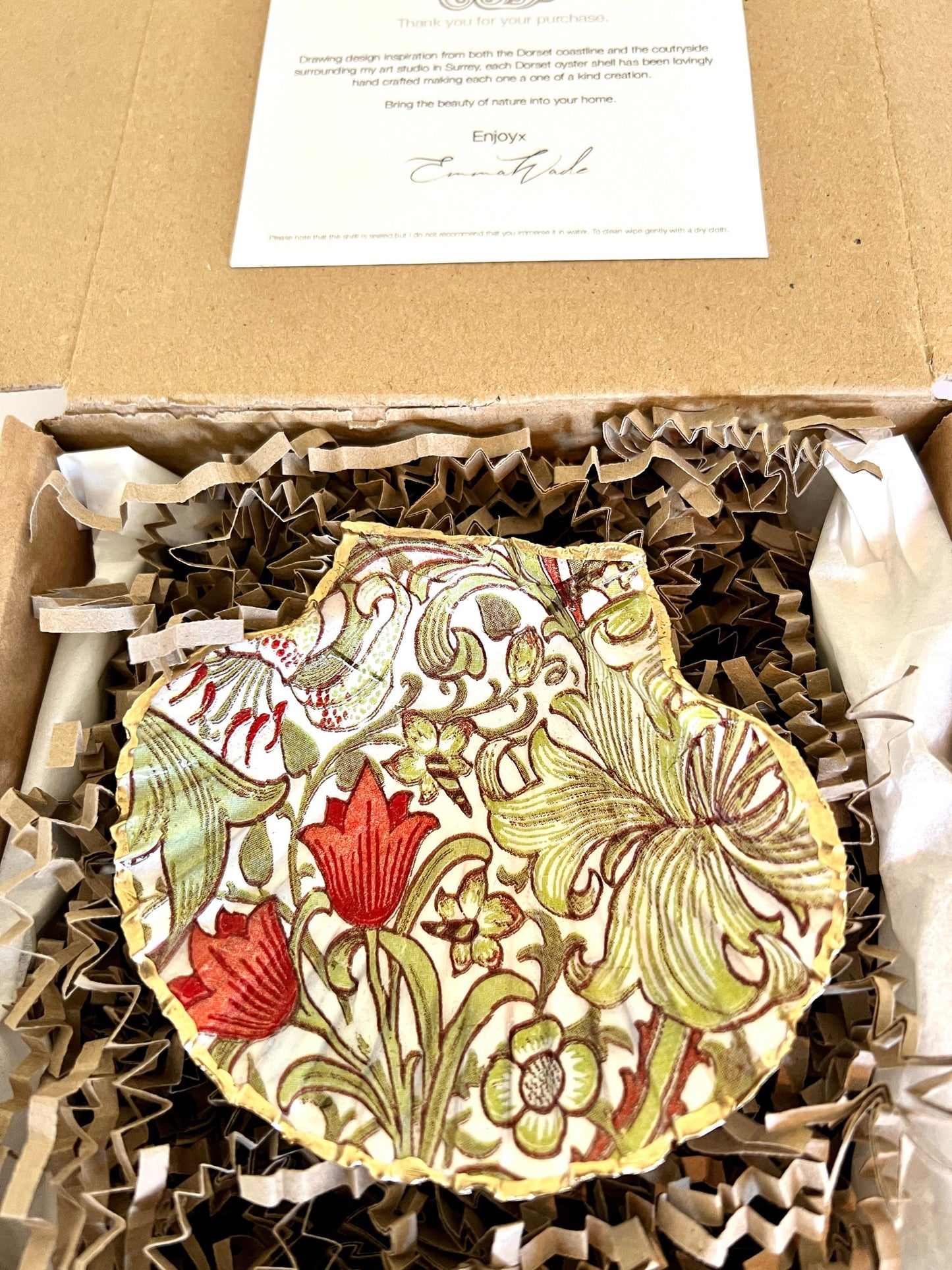 Handcrafted William Morris Golden Lily scallop shell ring holder featuring an elegant botanical floral pattern in green, cream and muted red tones. Decorative Dorset shell trinket dish finished with hand-painted gold leaf edging and sealed in glossy crystal resin for a luxurious shine. Coastal inspired jewellery tray and bedside catchall perfect for rings, earrings and keepsakes. Botanical shell decor for elegant interiors, cottage style homes, thoughtful handmade gifting.