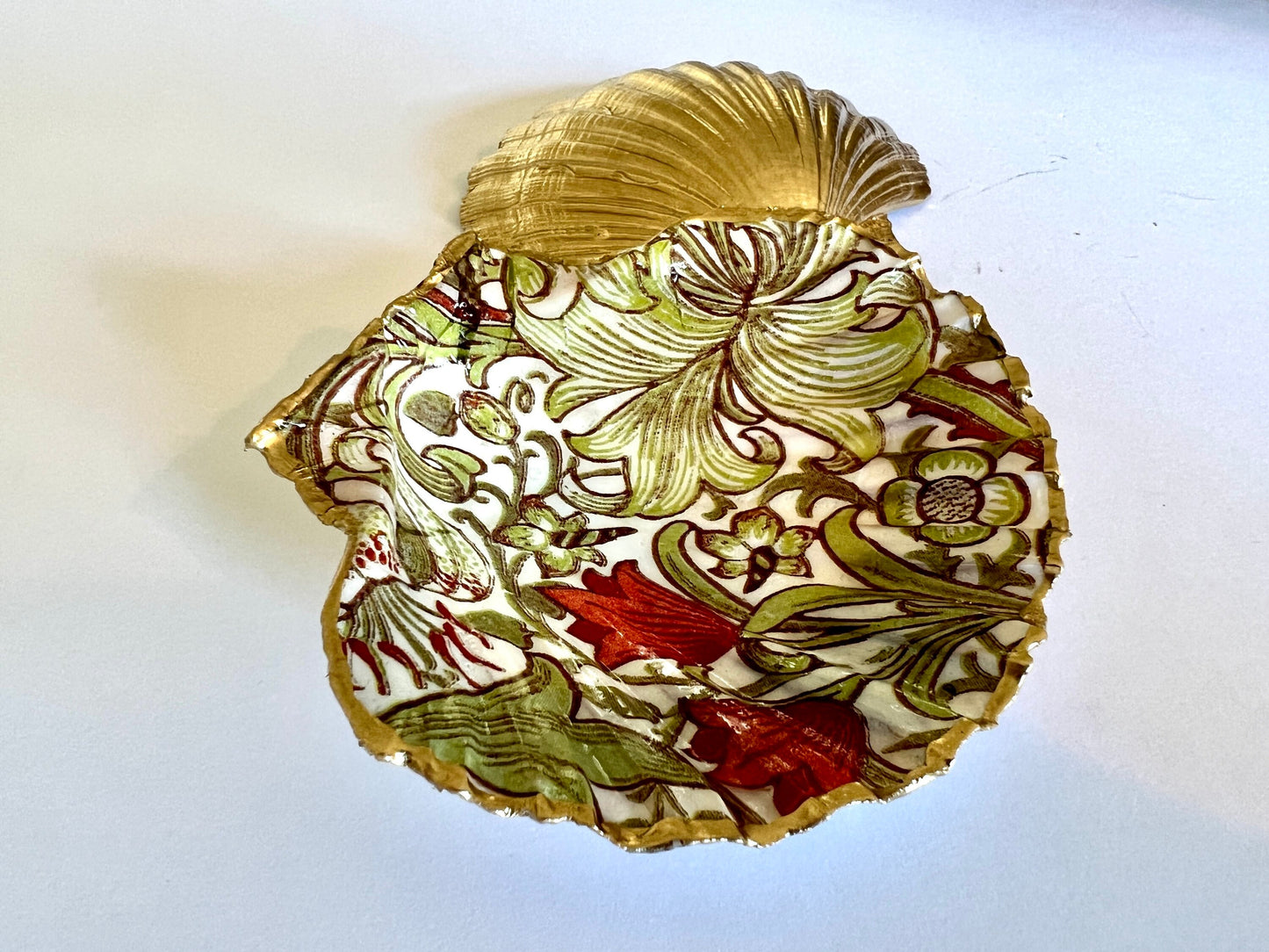 Handcrafted William Morris Golden Lily scallop shell ring holder featuring an elegant botanical floral pattern in green, cream and muted red tones. Decorative Dorset shell trinket dish finished with hand-painted gold leaf edging and sealed in glossy crystal resin for a luxurious shine. Coastal inspired jewellery tray and bedside catchall perfect for rings, earrings and keepsakes. Botanical shell decor for elegant interiors, cottage style homes, thoughtful handmade gifting.
