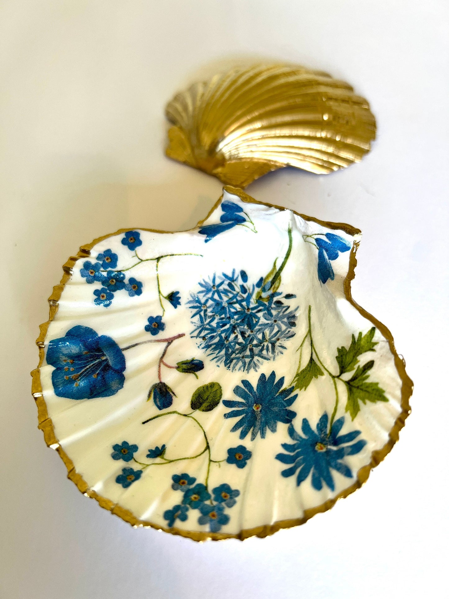 Handcrafted Dorset scallop shell trinket dish with blue spring flower design, finished with a gold leaf rim and glossy resin. The shell shows delicate blue floral motifs with green stems on a natural shell, styled on a light background. A coastal inspired jewellery or ring holder, perfect as a decorative keepsake dish, bathroom accent, or thoughtful handmade gift.