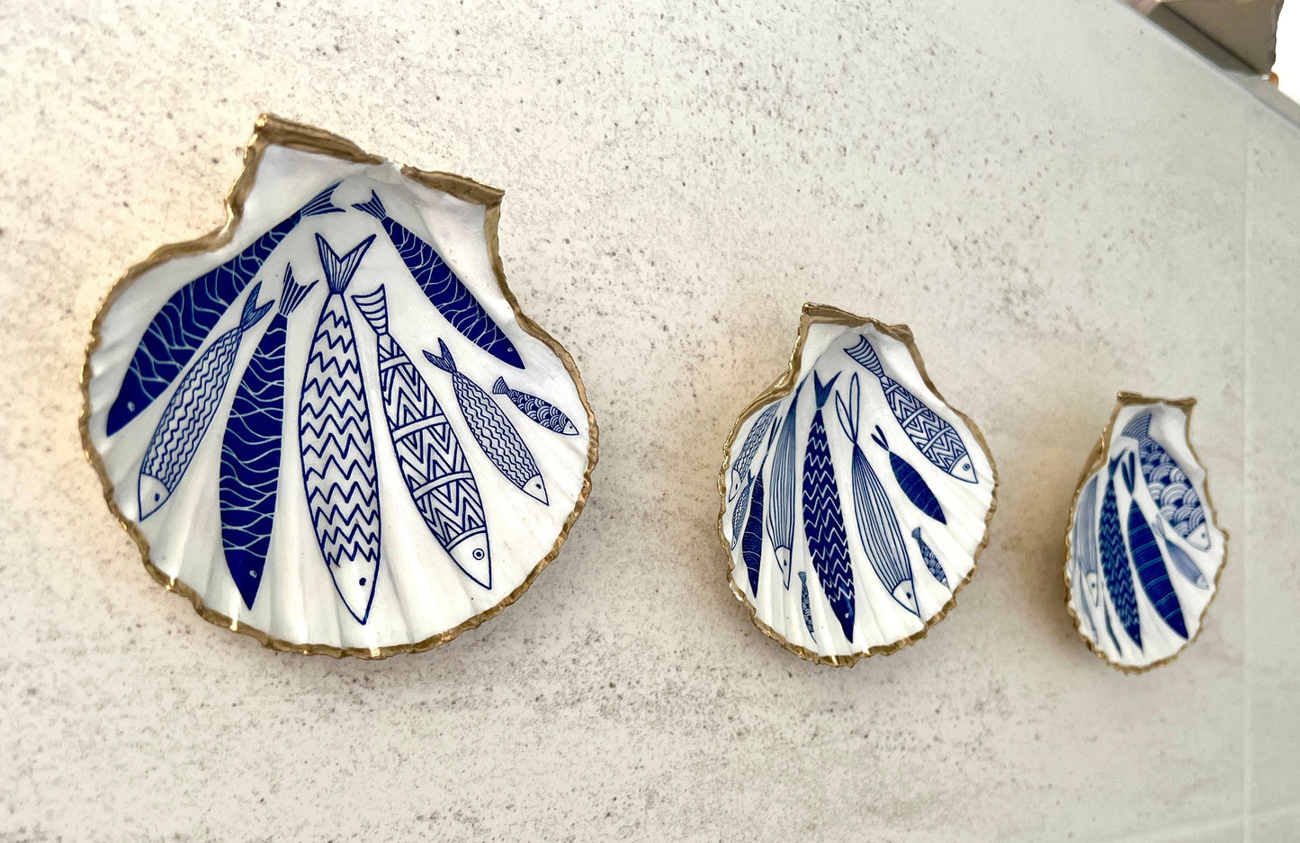 Handmade Dorset scallop shell trinket dishes featuring elegant blue fish illustrations with hand-painted gold leaf edging and glossy crystal clear resin. Coastal nautical jewellery dishes inspired by Scandinavian ocean decor, styled on a neutral background with detailed blue fish artwork and luxurious gilded edges.