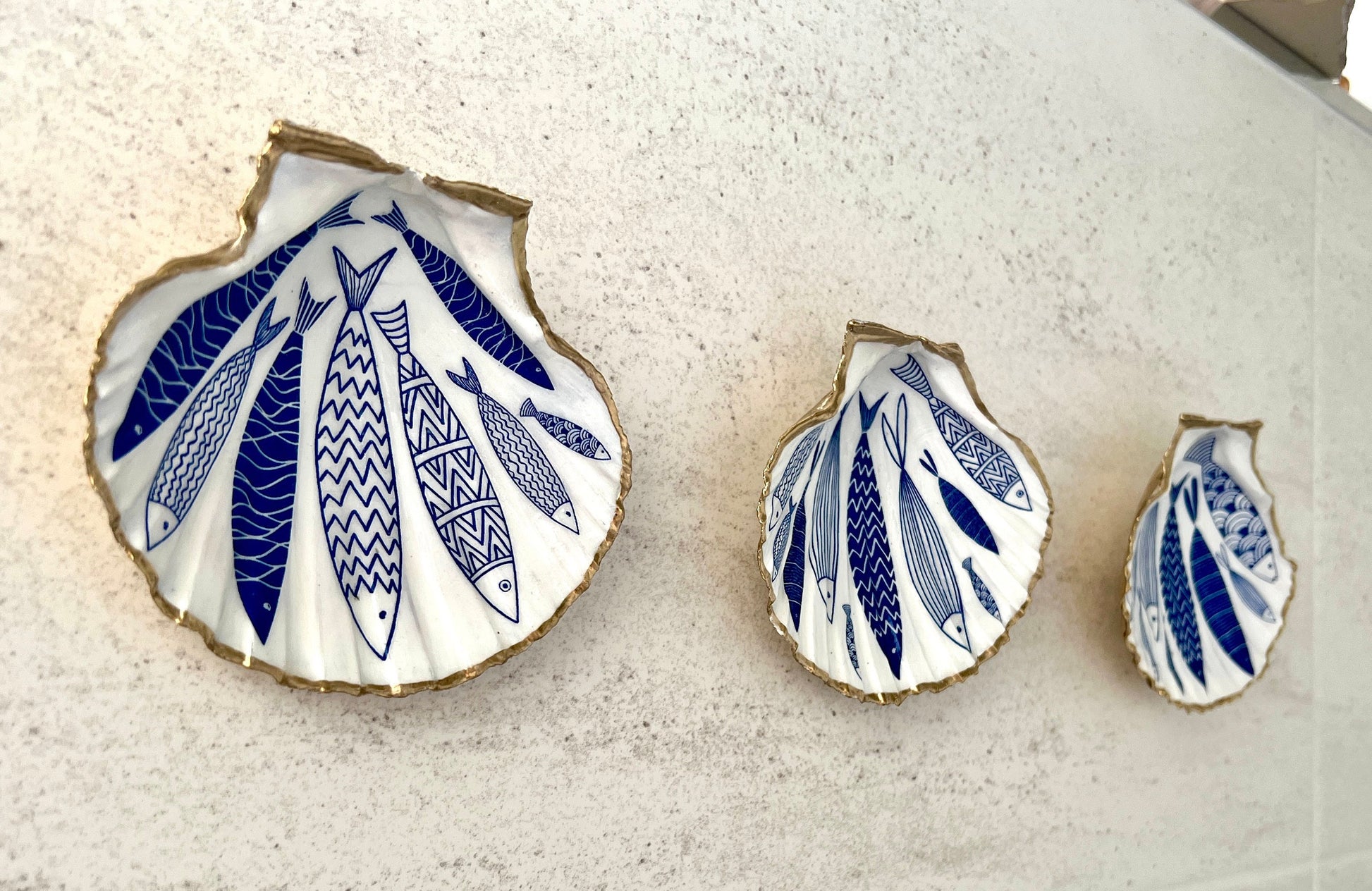 Handmade Dorset scallop shell trinket dishes featuring elegant blue fish illustrations with hand-painted gold leaf edging and glossy crystal clear resin. Coastal nautical jewellery dishes inspired by Scandinavian ocean decor, styled on a neutral background with detailed blue fish artwork and luxurious gilded edges.