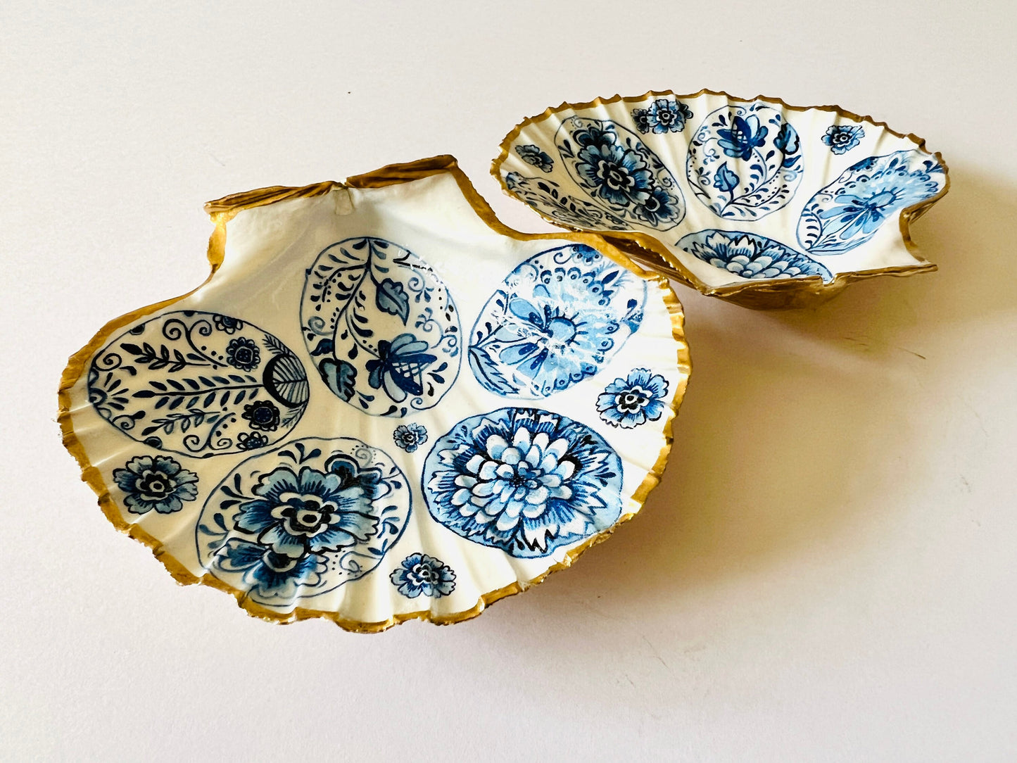 Handmade Dorset scallop shell trinket dishes featuring vintage inspired blue and white egg artwork with hand-painted gold leaf edging and glossy crystal clear resin. Elegant coastal jewellery dishes styled with Nordic and Scandinavian inspired porcelain patterns, photographed on soft neutral backgrounds with luxurious gilded detailing.