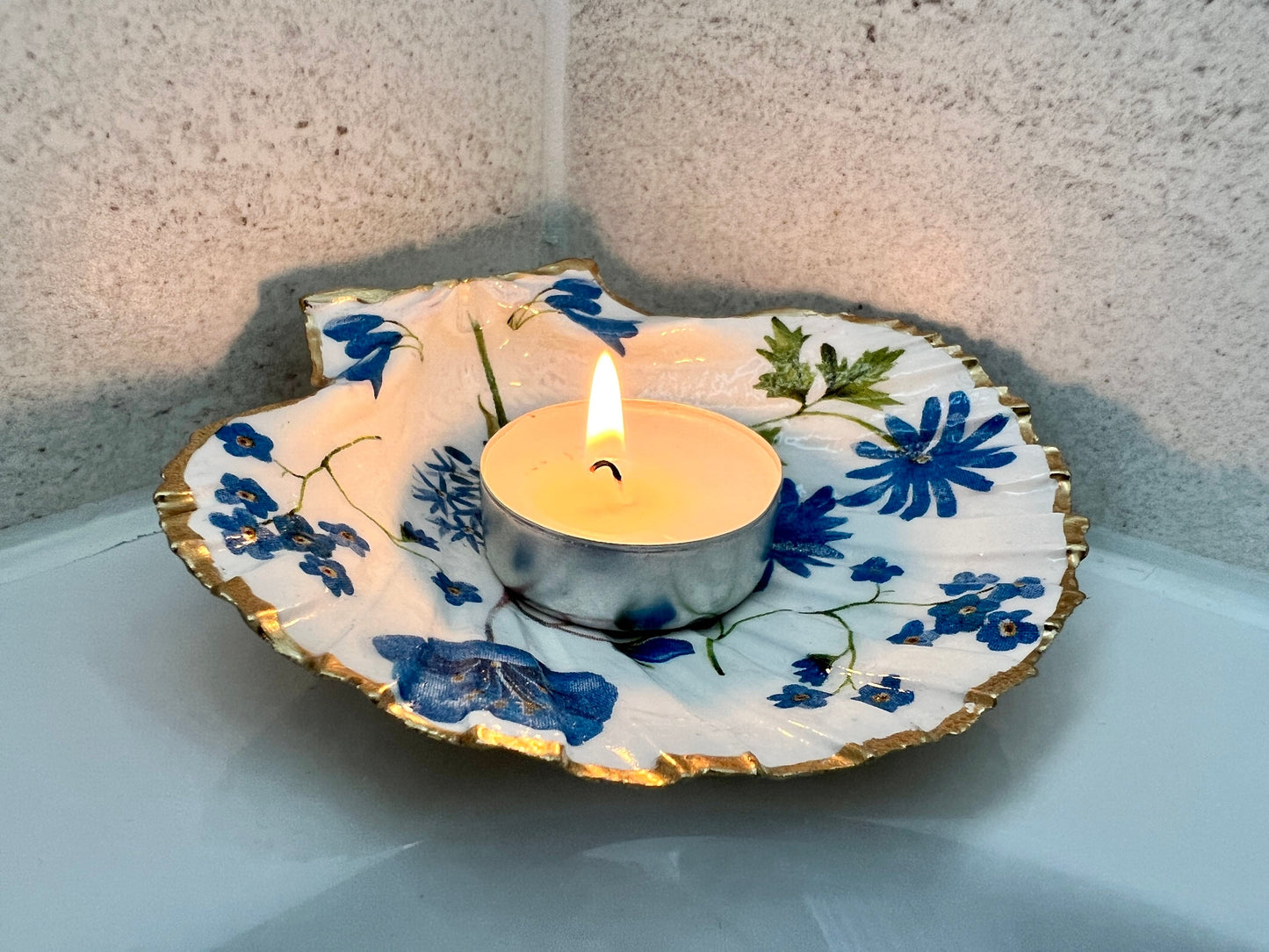 Handcrafted Dorset scallop shell trinket dish with blue spring flower design, finished with a gold leaf rim and glossy resin. The shell shows delicate blue floral motifs with green stems on a natural shell, styled on a light background. A coastal inspired jewellery or ring holder, perfect as a decorative keepsake dish, bathroom accent, or thoughtful handmade gift.