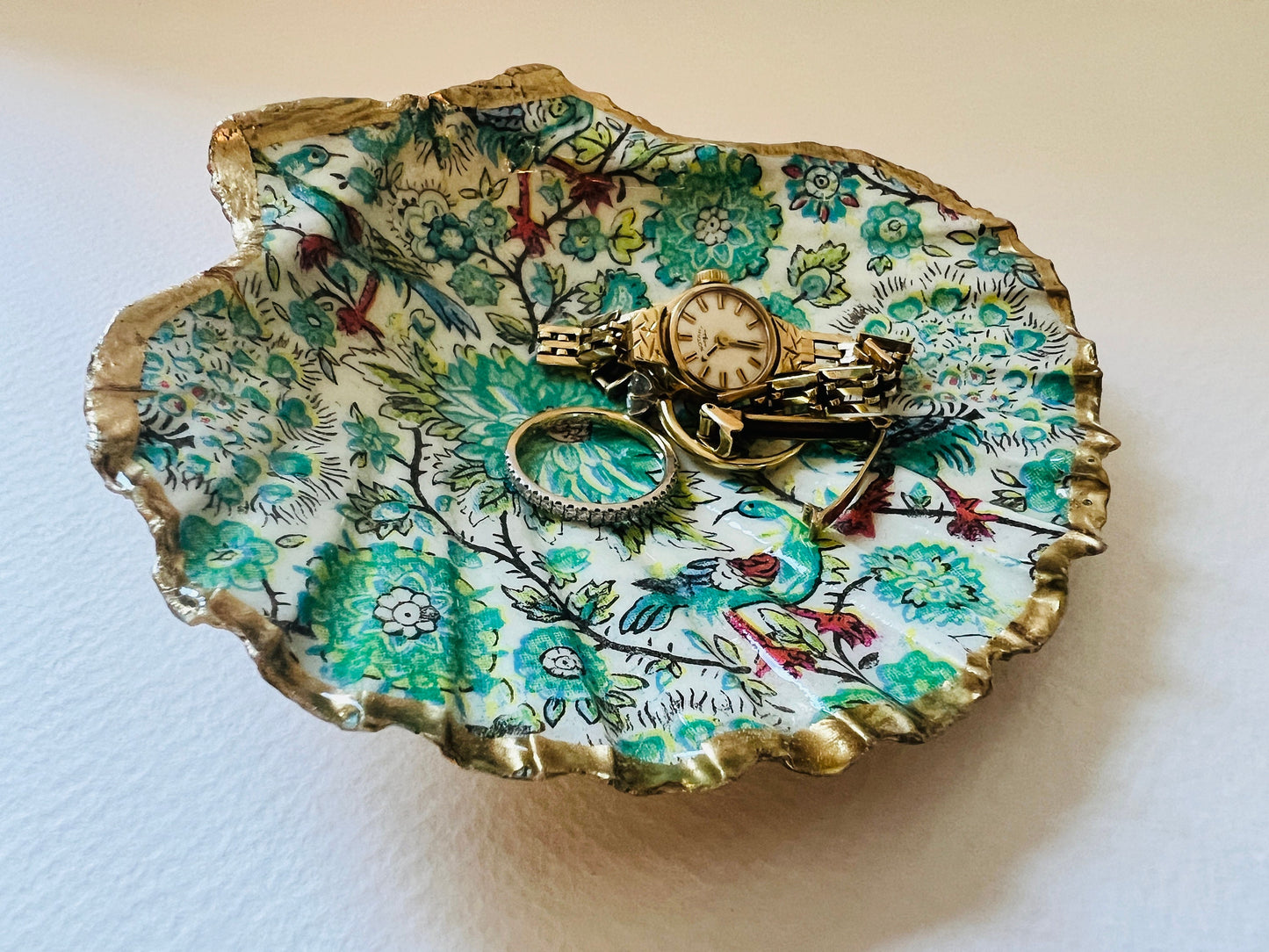 Handmade Dorset scallop shell trinket dishes featuring intricate green peacock inspired artwork in emerald, turquoise and gold tones with hand-painted gold leaf edging and glossy crystal clear resin. Elegant coastal jewellery dishes styled on a neutral background with nature inspired bird motifs and luxurious gilded detailing.