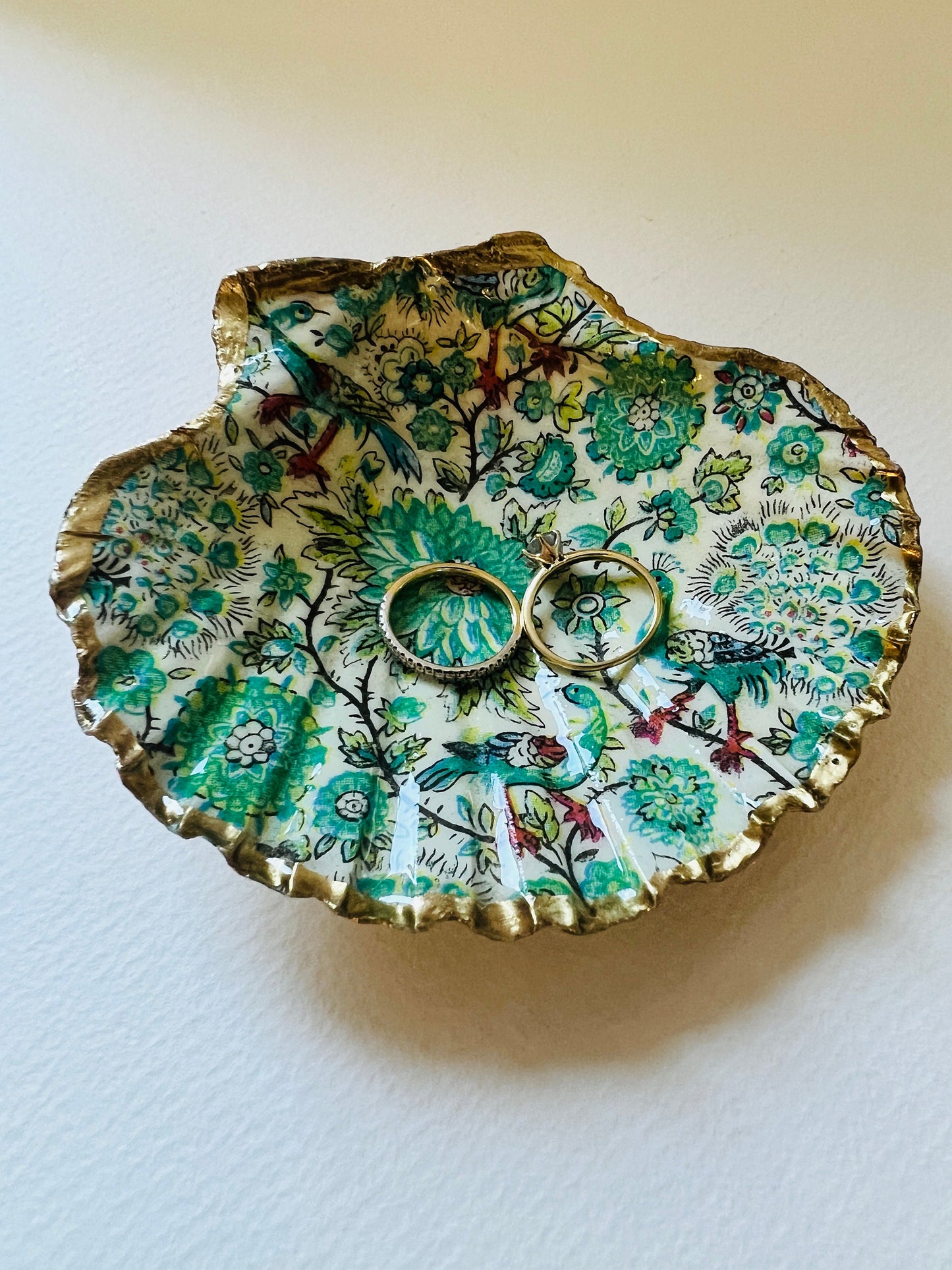 Handmade Dorset scallop shell trinket dishes featuring intricate green peacock inspired artwork in emerald, turquoise and gold tones with hand-painted gold leaf edging and glossy crystal clear resin. Elegant coastal jewellery dishes styled on a neutral background with nature inspired bird motifs and luxurious gilded detailing.