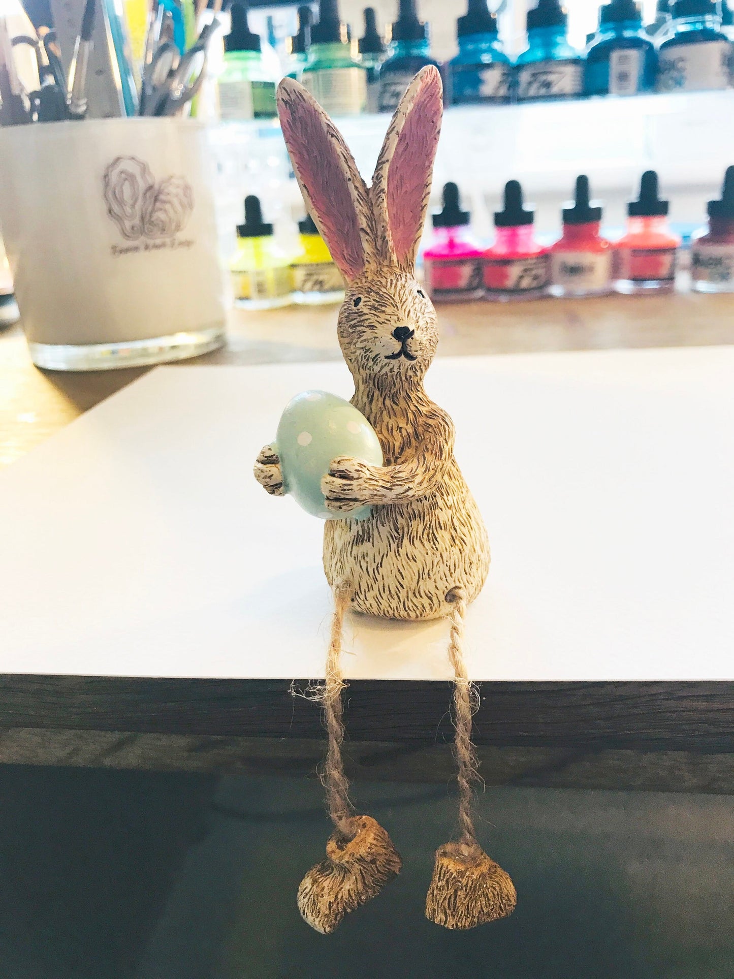 Hand-finished shelf sitting bunny ornament with dangling jute legs and polka dot egg. Available in pink or green. Perfect Easter rabbit shelf decoration, birthday gift, or home décor accessory. Cute animal figurine for rabbit lovers, Easter decorations, new home presents, or seasonal mantel styling.