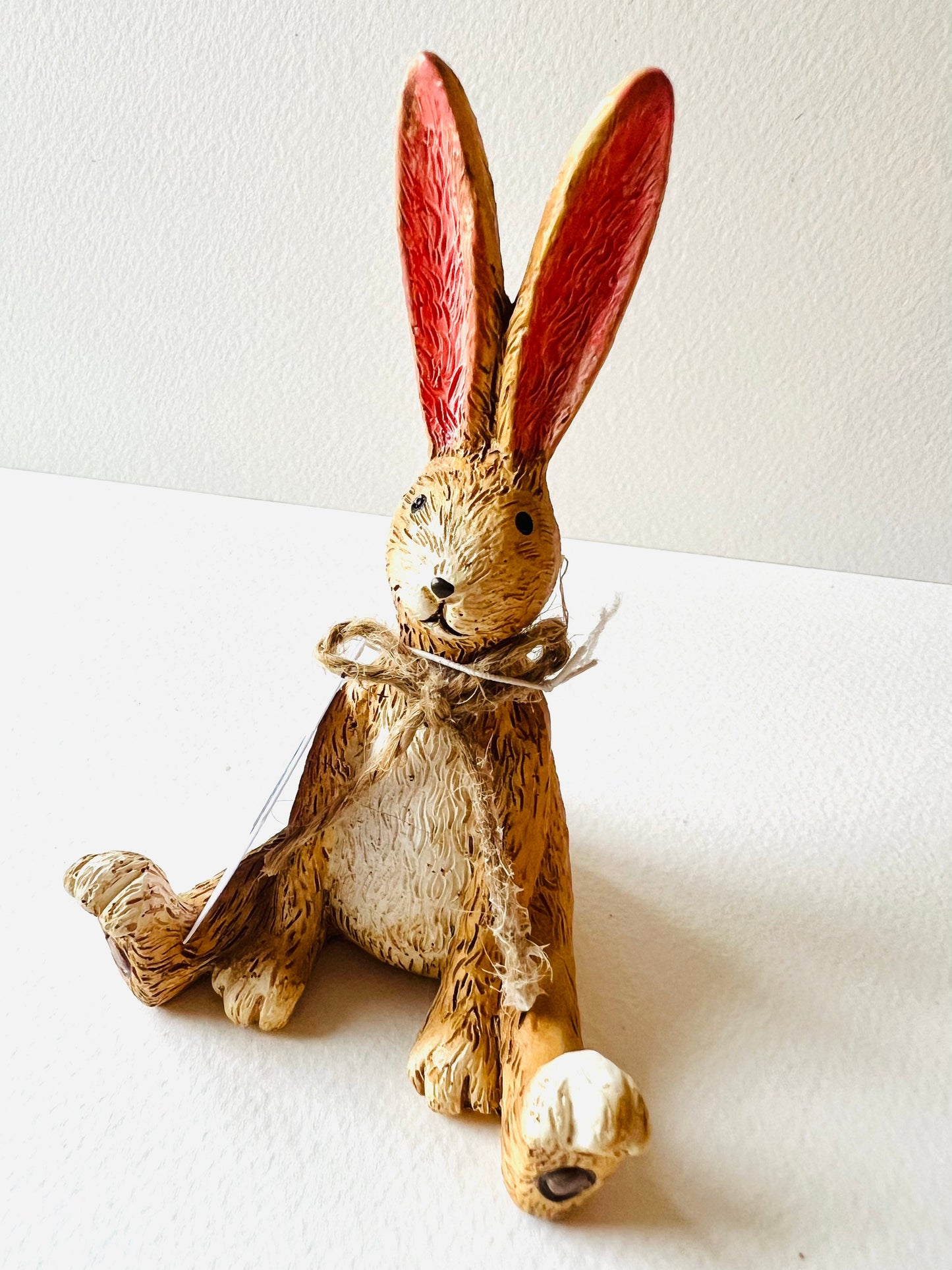 Hand finished polyresin sitting bunny ornament with upright pink ears and rustic jute bow, textured brown rabbit figurine displayed on wooden stand.