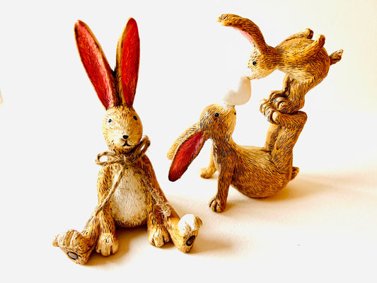 Set of two hand finished polyresin bunny ornaments, including seated rabbit with jute bow and rabbit with baby figure, rustic brown textured finish with pink ears, styled on neutral background as decorative shelf decor.