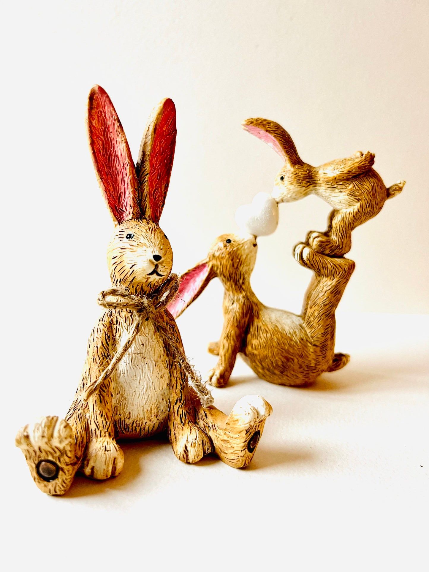 Hand finished polyresin Christmas bunny ornaments with rustic jute bows and soft pink inner ears. Decorative rabbit shelf ornaments shown as a seated bunny and two bunnies kissing, available individually or as a set. Festive woodland-style decor for Christmas shelves, mantelpieces, cottage interiors and rabbit lover gifts.