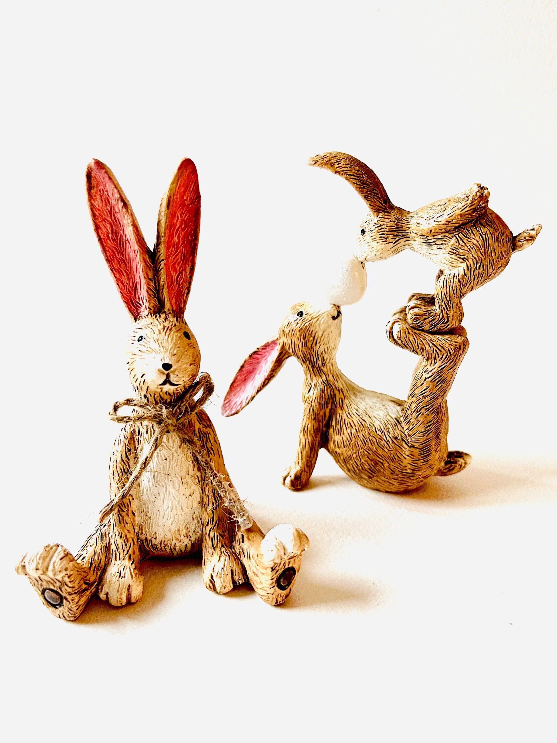 Set of two hand finished polyresin bunny ornaments, including seated rabbit with jute bow and rabbit with baby figure, rustic brown textured finish with pink ears, styled on neutral background as decorative shelf decor.