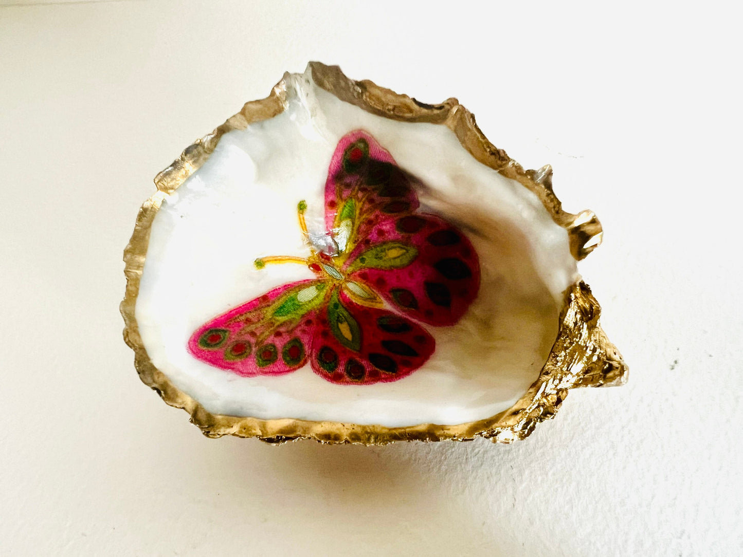 Handmade Dorset oyster shell ring dishes featuring colourful butterfly designs in green, blue and pink tones. Decorative oyster shell trinket holders finished with hand-applied liquid gold leaf edging and glossy resin. Nature inspired jewellery dishes with natural pearlescent shell interiors, styled as coastal décor, ring holders and keepsake gifts.