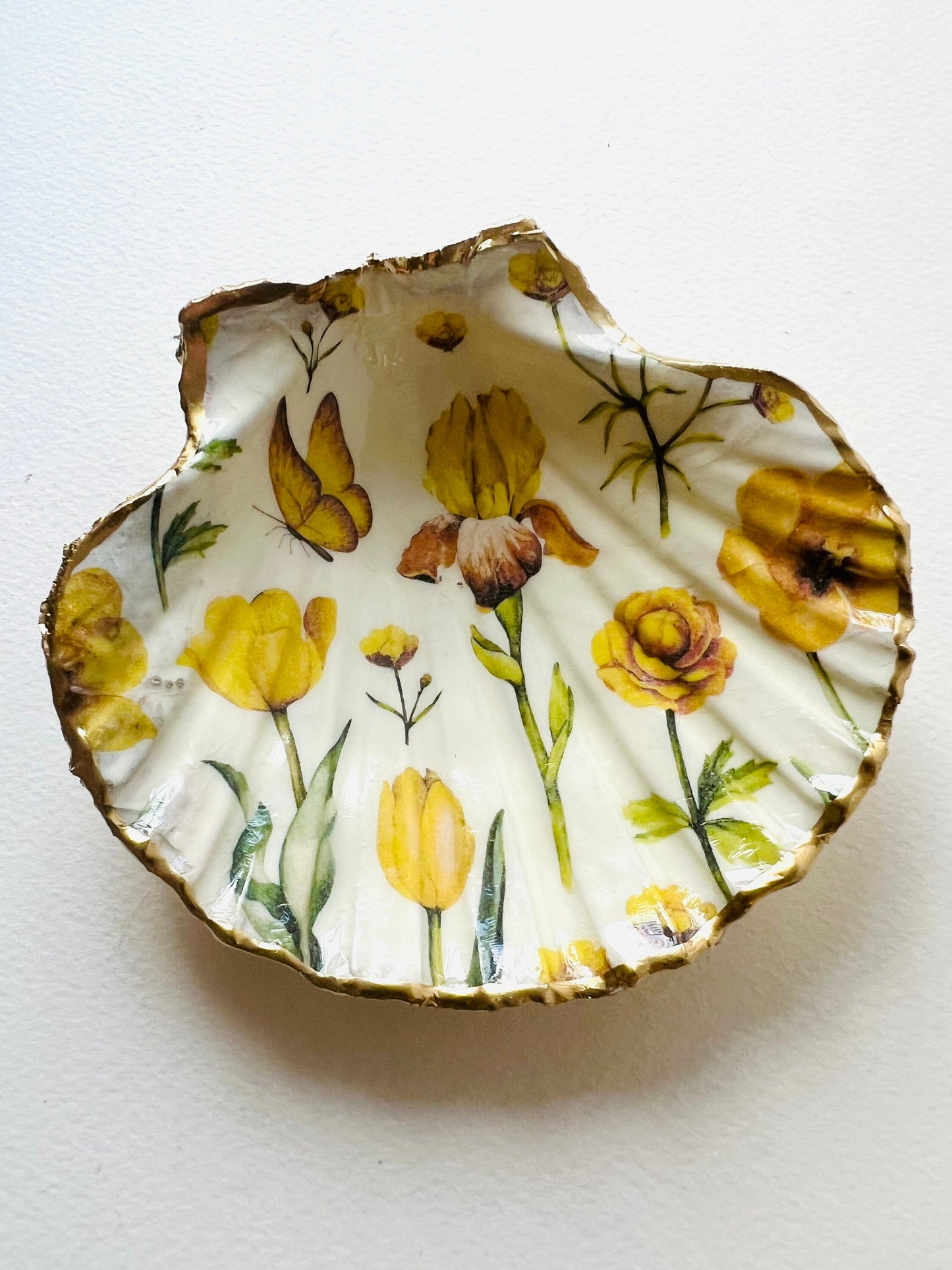 Hand finished scallop shell trinket dish featuring yellow spring flower artwork with a hand gilded gold leaf rim, sealed in glossy clear resin for durability and shine. Coastal inspired shell jewellery dish photographed on a light surface, ideal for rings, earrings and small keepsakes. Decorative floral shell dish by EmmaWadeART inspired by the Dorset coastline, perfect as a gift or elegant home décor accent.