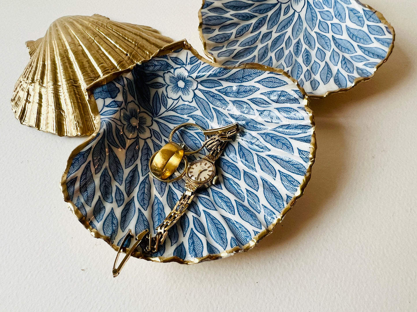 Handmade Dorset scallop shell trinket dishes featuring intricate blue floral and leaf artwork with hand-painted gold leaf edging and glossy crystal clear resin. Coastal botanical jewellery dishes inspired by Nordic Scandinavian decor, styled on a neutral background with elegant blue flower detailing and gilded edges.