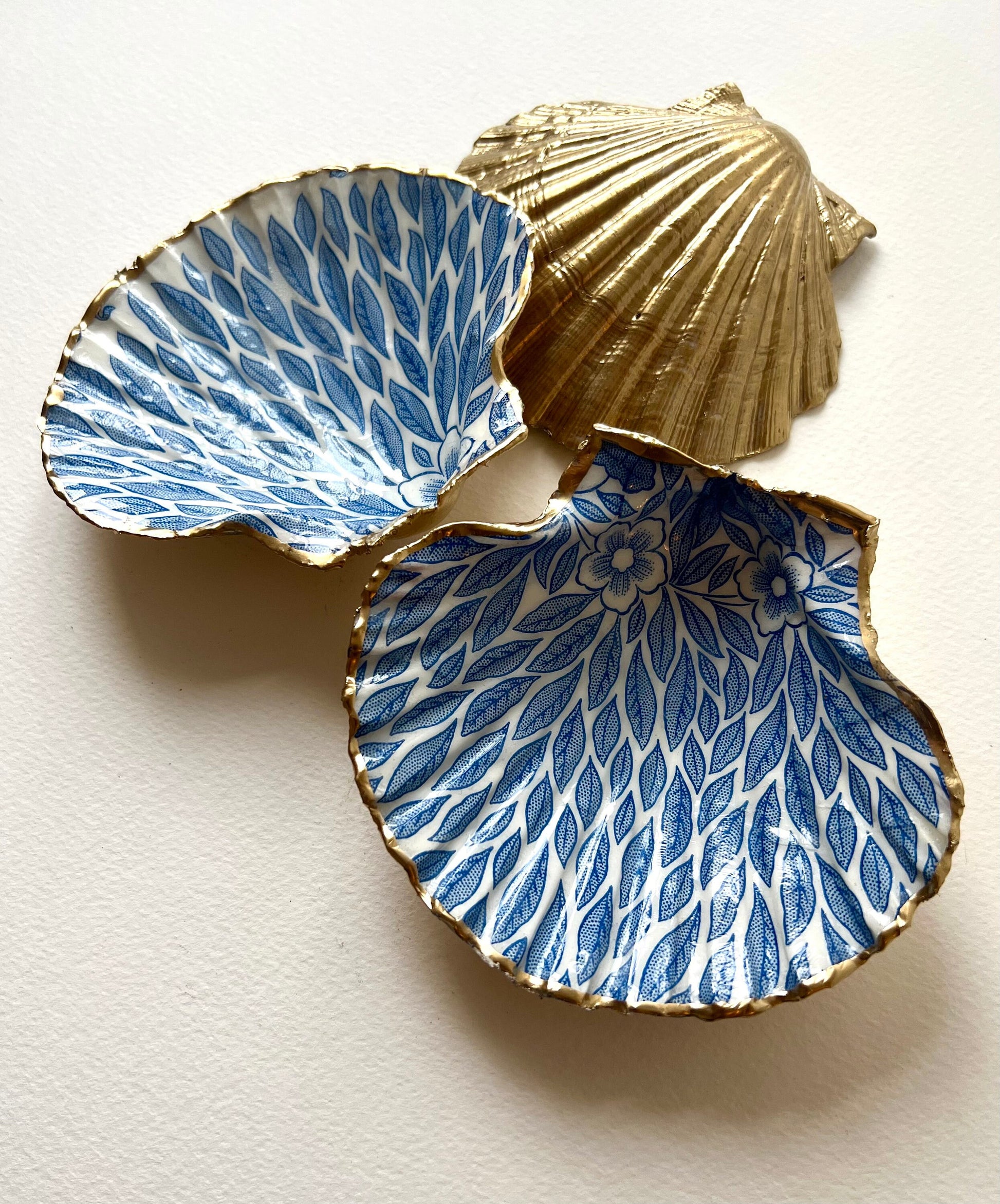 Handmade Dorset scallop shell trinket dishes featuring intricate blue floral and leaf artwork with hand-painted gold leaf edging and glossy crystal clear resin. Coastal botanical jewellery dishes inspired by Nordic Scandinavian decor, styled on a neutral background with elegant blue flower detailing and gilded edges.