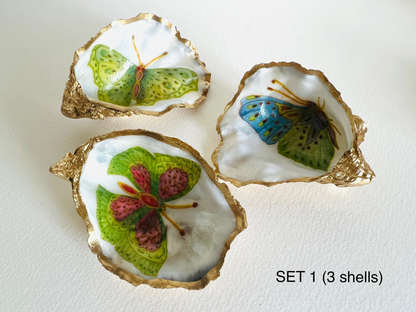 Handmade Dorset oyster shell ring dishes featuring colourful butterfly designs in green, blue and pink tones. Decorative oyster shell trinket holders finished with hand-applied liquid gold leaf edging and glossy resin. Nature inspired jewellery dishes with natural pearlescent shell interiors, styled as coastal décor, ring holders and keepsake gifts.