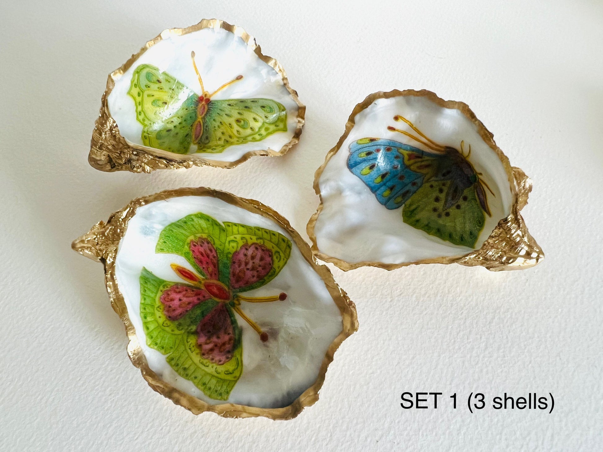 Handmade Dorset oyster shell ring dishes featuring colourful butterfly designs in green, blue and pink tones. Decorative oyster shell trinket holders finished with hand-applied liquid gold leaf edging and glossy resin. Nature inspired jewellery dishes with natural pearlescent shell interiors, styled as coastal décor, ring holders and keepsake gifts.