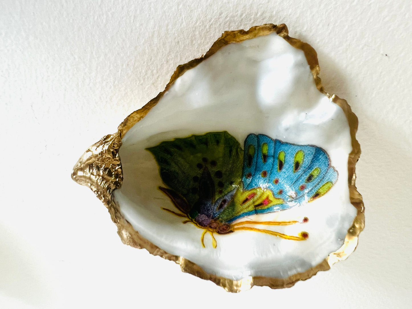 Handmade Dorset oyster shell ring dishes featuring colourful butterfly designs in green, blue and pink tones. Decorative oyster shell trinket holders finished with hand-applied liquid gold leaf edging and glossy resin. Nature inspired jewellery dishes with natural pearlescent shell interiors, styled as coastal décor, ring holders and keepsake gifts.