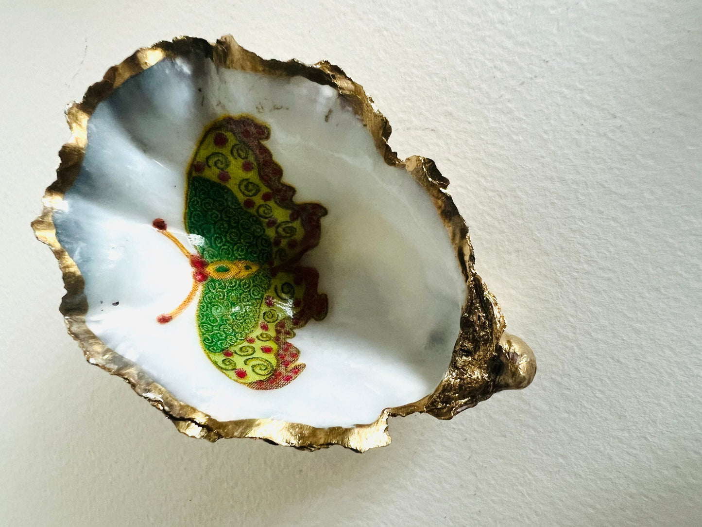 Handmade Dorset oyster shell ring dishes featuring colourful butterfly designs in green, blue and pink tones. Decorative oyster shell trinket holders finished with hand-applied liquid gold leaf edging and glossy resin. Nature inspired jewellery dishes with natural pearlescent shell interiors, styled as coastal décor, ring holders and keepsake gifts.
