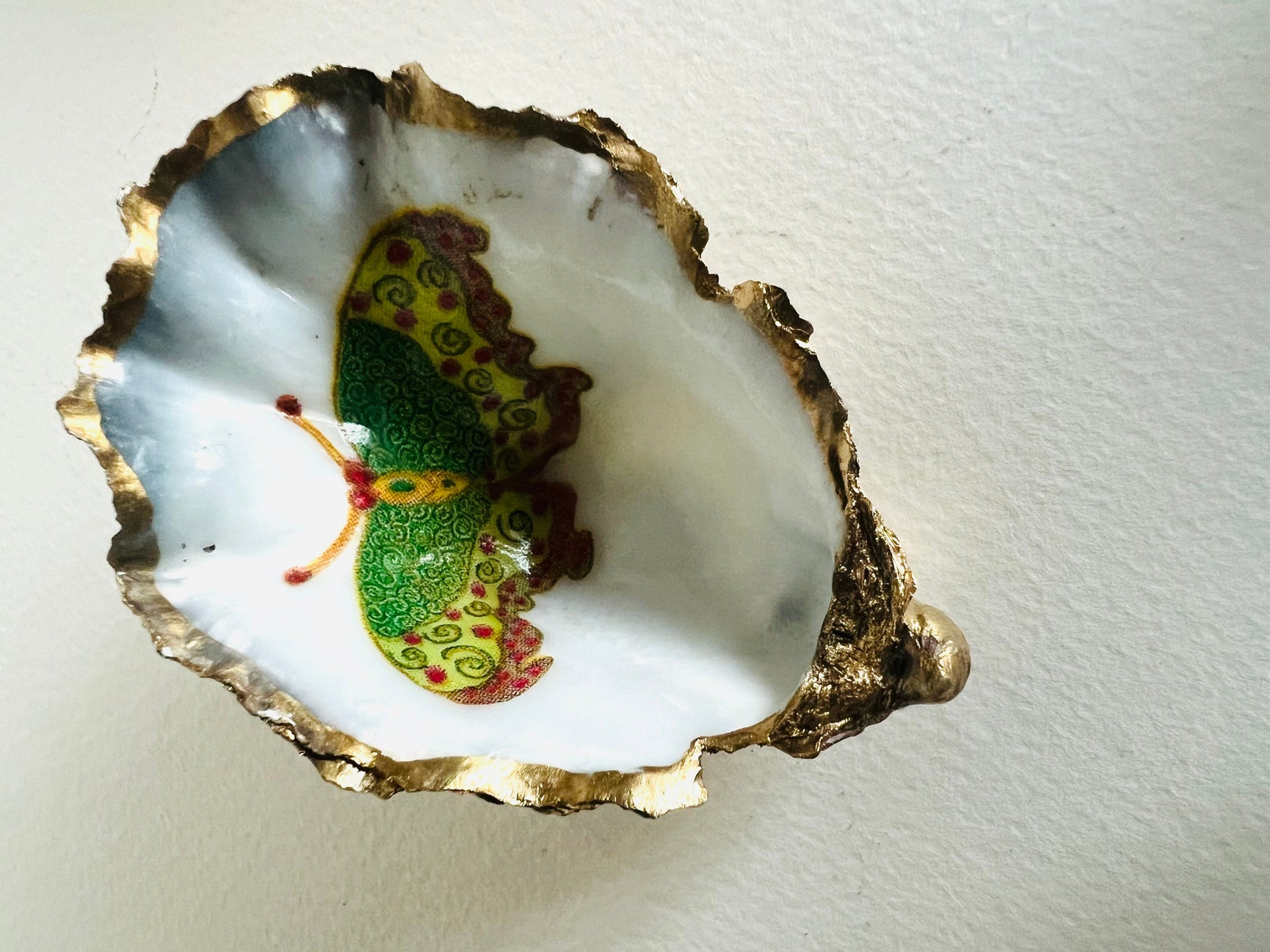 Handmade Dorset oyster shell ring dishes featuring colourful butterfly designs in green, blue and pink tones. Decorative oyster shell trinket holders finished with hand-applied liquid gold leaf edging and glossy resin. Nature inspired jewellery dishes with natural pearlescent shell interiors, styled as coastal décor, ring holders and keepsake gifts.