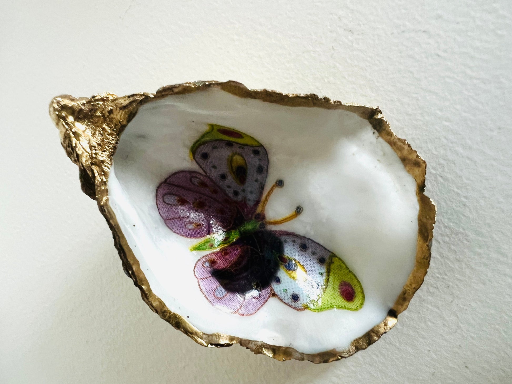 Handmade Dorset oyster shell ring dishes featuring colourful butterfly designs in green, blue and pink tones. Decorative oyster shell trinket holders finished with hand-applied liquid gold leaf edging and glossy resin. Nature inspired jewellery dishes with natural pearlescent shell interiors, styled as coastal décor, ring holders and keepsake gifts.