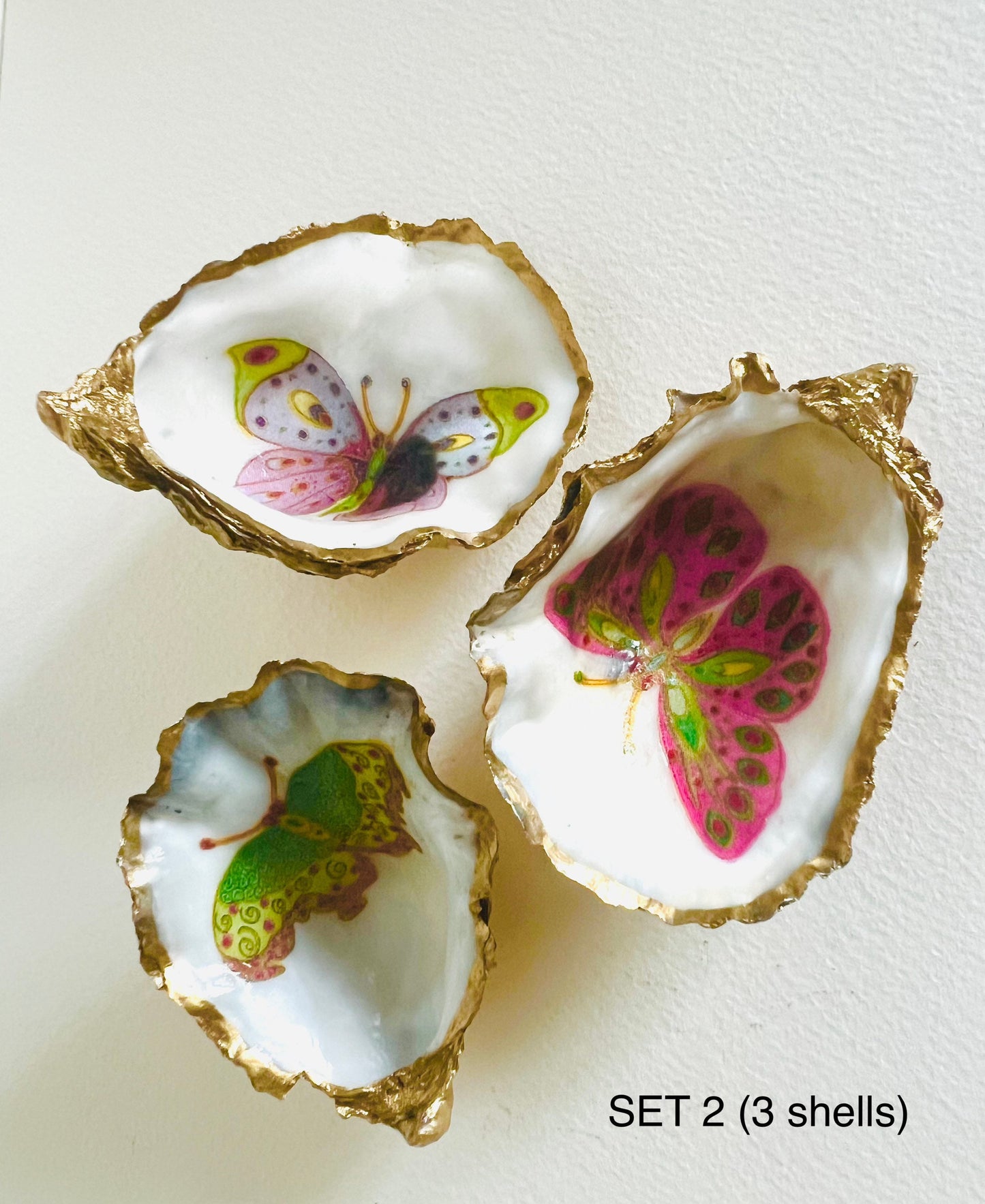 Handmade Dorset oyster shell ring dishes featuring colourful butterfly designs in green, blue and pink tones. Decorative oyster shell trinket holders finished with hand-applied liquid gold leaf edging and glossy resin. Nature inspired jewellery dishes with natural pearlescent shell interiors, styled as coastal décor, ring holders and keepsake gifts.