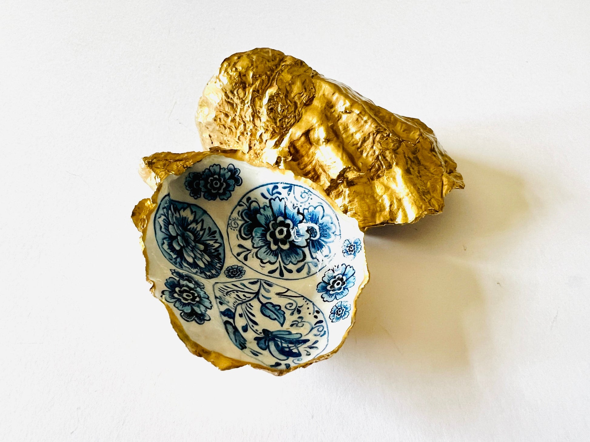 Handmade Dorset oyster shell trinket dish featuring a vintage blue and white decorative egg design with intricate floral porcelain style detailing. Oyster shell is hand gilded with liquid gold leaf edging and sealed in glossy crystal clear resin. Elegant coastal jewelry holder, ring dish, trinket tray, or decorative keepsake inspired by antique ceramics and vintage decor styling. Luxury handmade shell art photographed on a marble background.