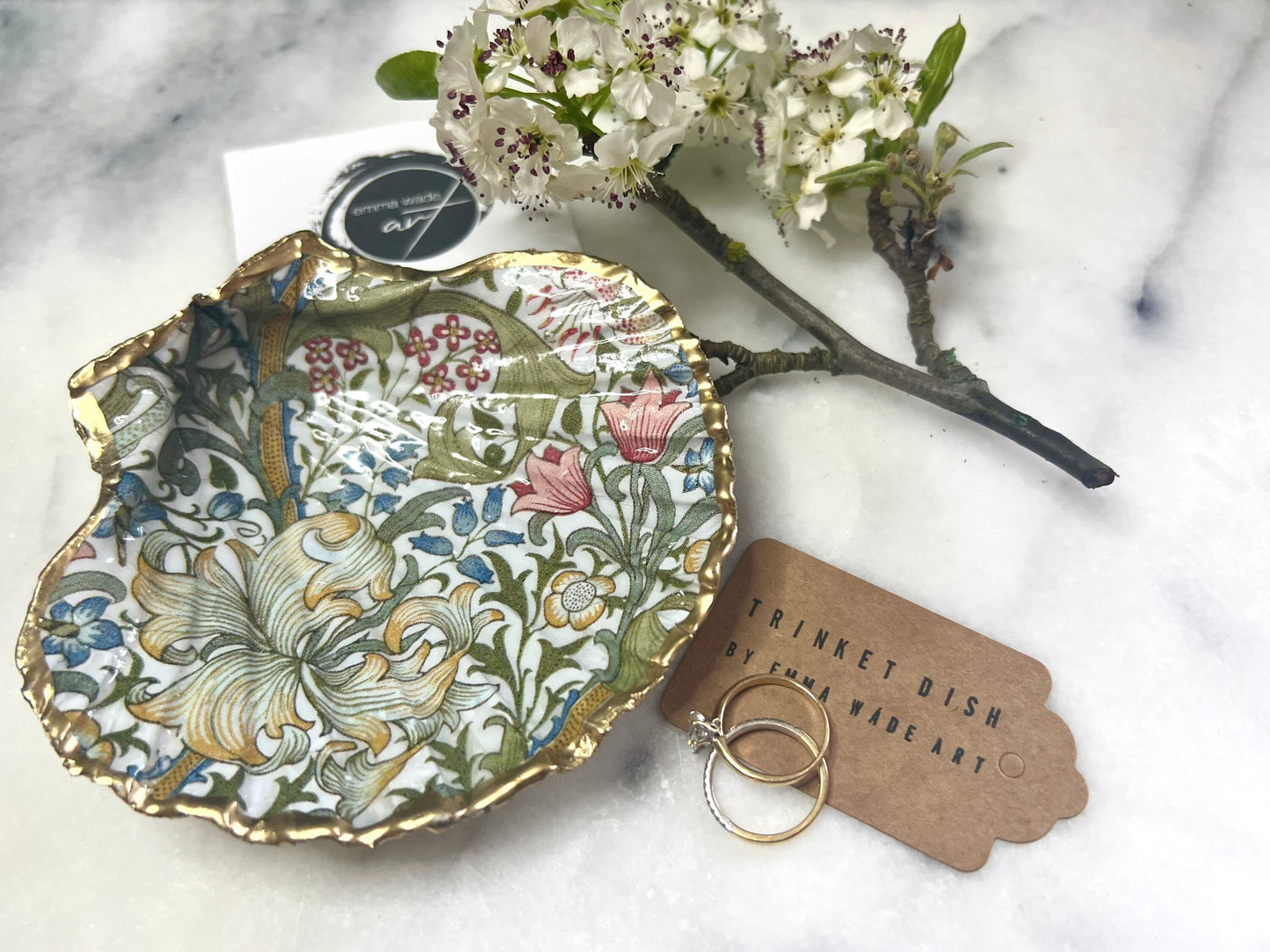 Handcrafted Dorset scallop shell trinket dish featuring William Morris Golden Lily floral design. Natural shell edged with hand applied gold leaf and sealed with glossy resin. Heritage floral jewellery holder displayed with rings on marble surface. Elegant coastal home decor gift.
