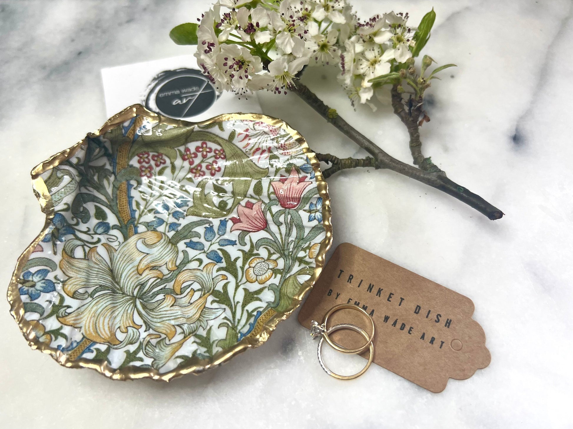 Handcrafted Dorset scallop shell trinket dish featuring William Morris Golden Lily floral design. Natural shell edged with hand applied gold leaf and sealed with glossy resin. Heritage floral jewellery holder displayed with rings on marble surface. Elegant coastal home decor gift.