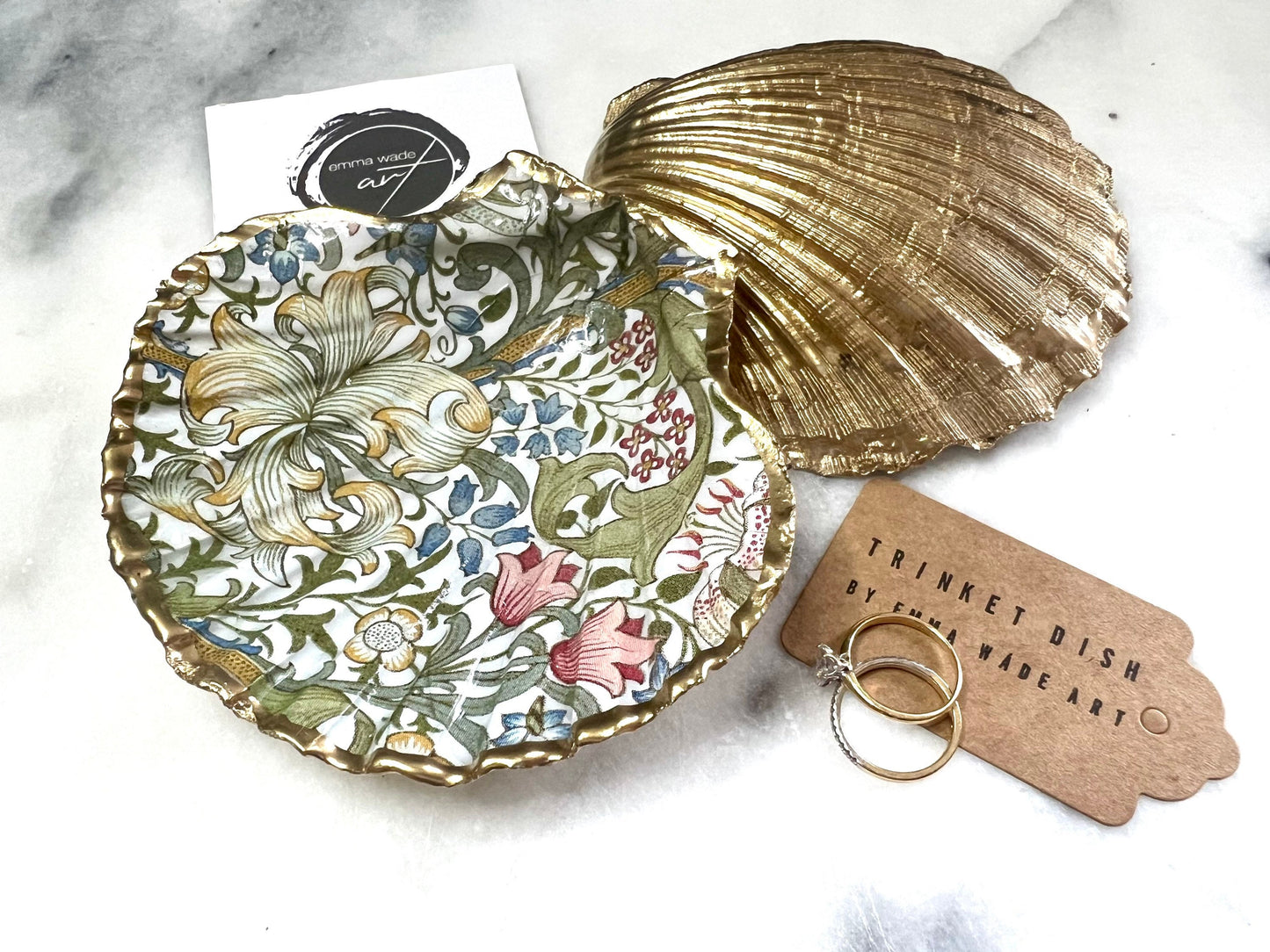 Handcrafted Dorset scallop shell trinket dish featuring William Morris Golden Lily floral design. Natural shell edged with hand applied gold leaf and sealed with glossy resin. Heritage floral jewellery holder displayed with rings on marble surface. Elegant coastal home decor gift.