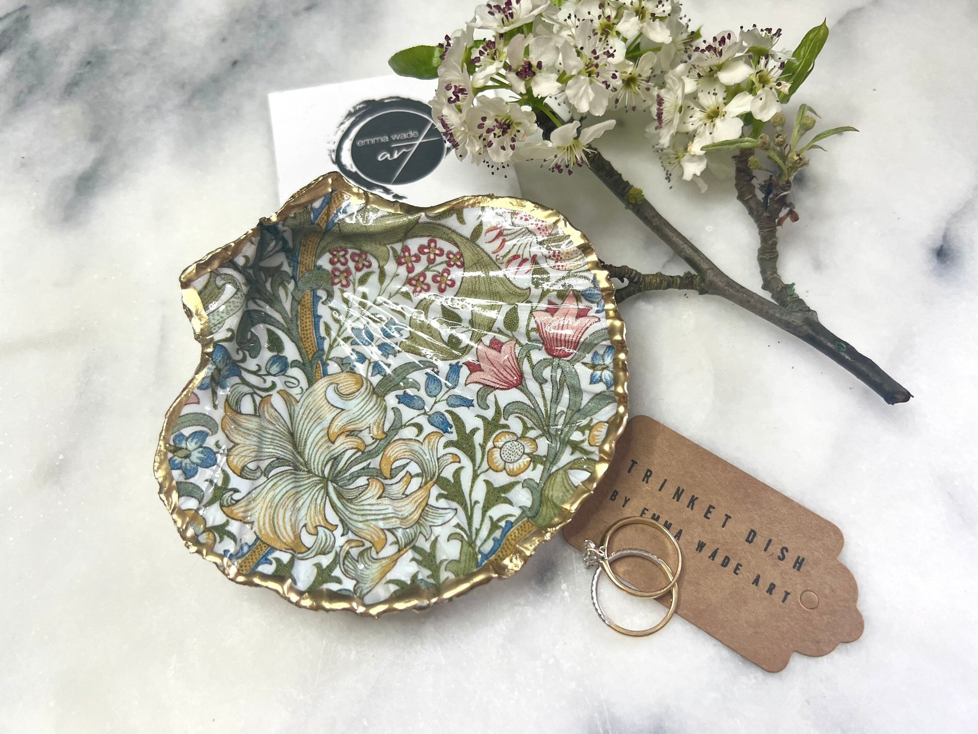 Handcrafted Dorset scallop shell trinket dish featuring William Morris Golden Lily floral design. Natural shell edged with hand applied gold leaf and sealed with glossy resin. Heritage floral jewellery holder displayed with rings on marble surface. Elegant coastal home decor gift.