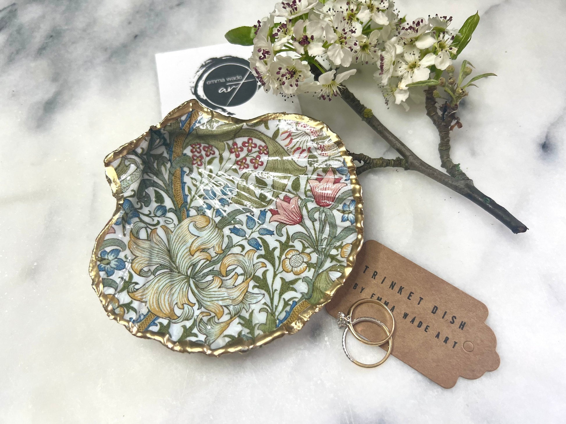 Handcrafted Dorset scallop shell trinket dish featuring William Morris Golden Lily floral design. Natural shell edged with hand applied gold leaf and sealed with glossy resin. Heritage floral jewellery holder displayed with rings on marble surface. Elegant coastal home decor gift.
