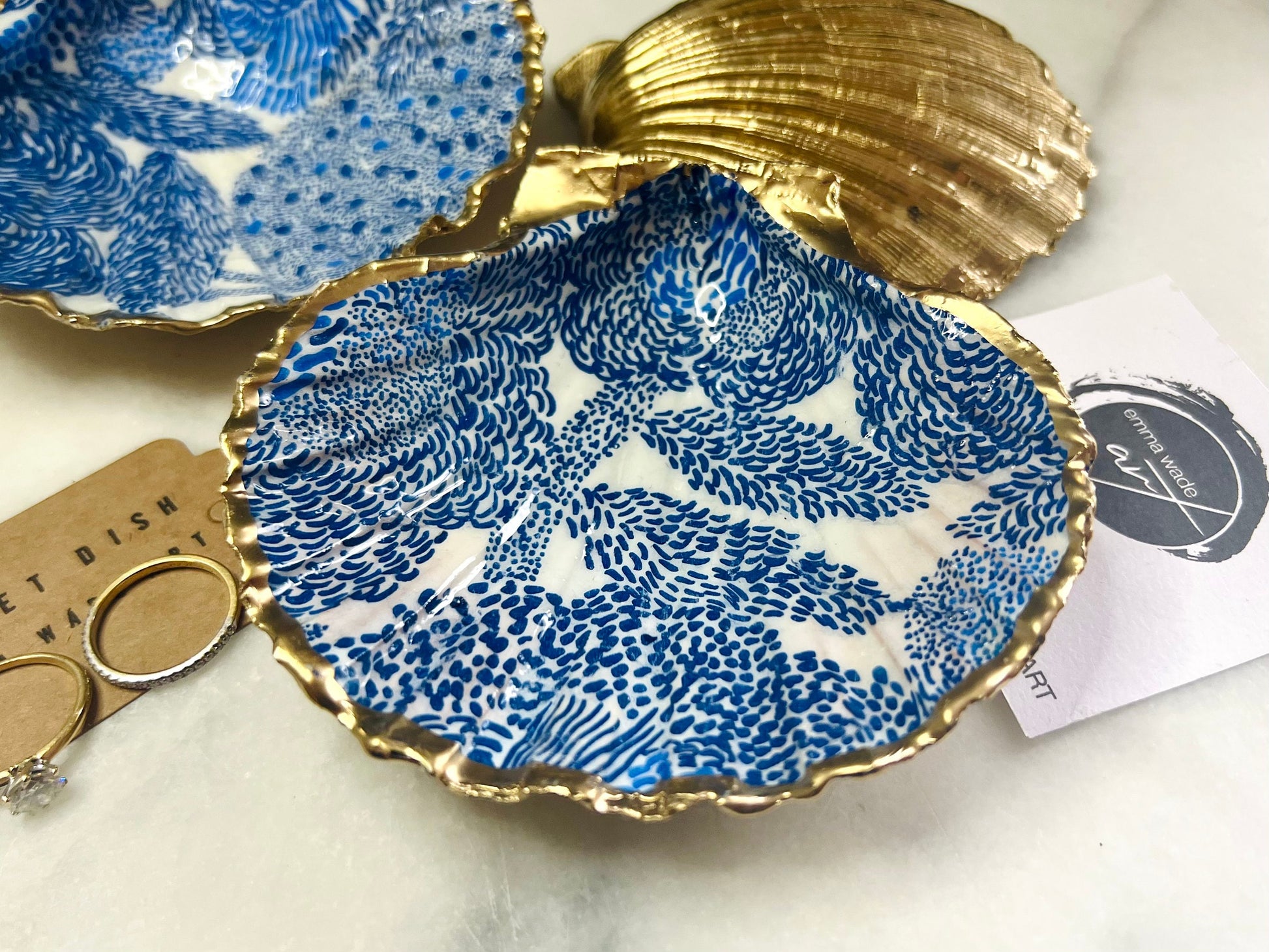 Marimekko inspired blue floral scallop shell trinket dish with hand-applied gold leaf edging and glossy resin finish. Natural Dorset shell jewellery holder featuring bold Scandinavian style botanical pattern. Decorative coastal ring dish styled on a marble surface, ideal as a bedside trinket bowl, host gift or luxury handmade home accent.