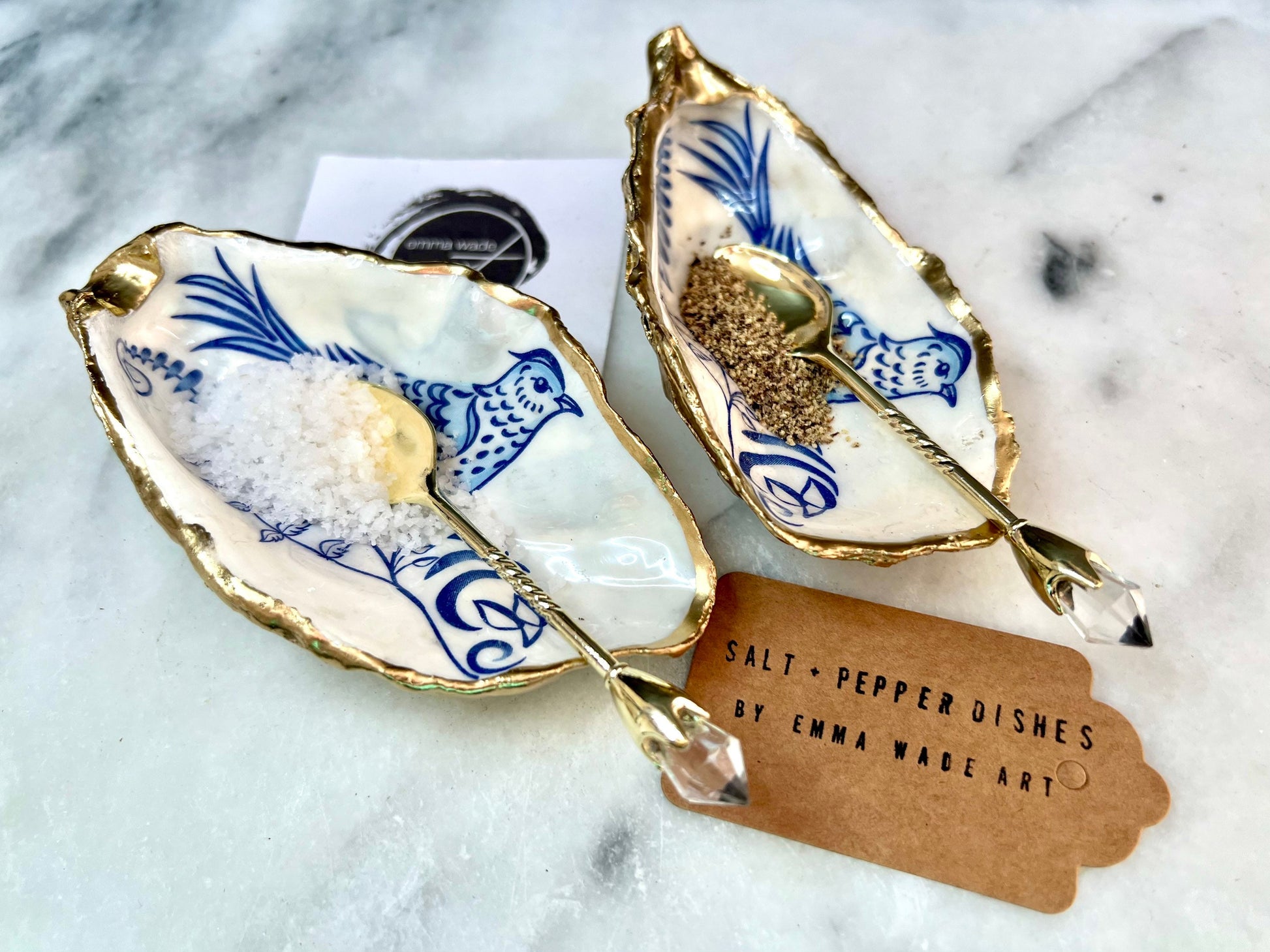 Handmade blue bird oyster shell salt and pepper pinch pot set crafted from natural Dorset oyster shells with glossy pearlescent interiors, blue porcelain style bird artwork and shimmering liquid gold leaf edging. Luxury coastal cruet dishes sealed with food-safe resin and styled with optional gold spoons. Elegant seaside dining decor for beach house kitchens, dinner parties, weddings, anniversaries and bird lovers. Artisan shell tableware handmade in the UK with coastal blue and white styling.