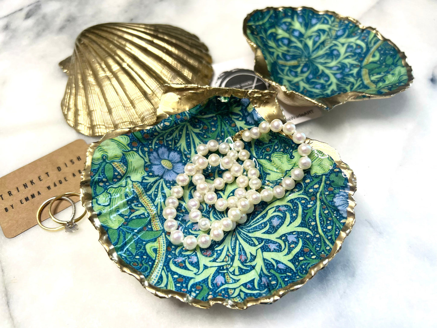 Handmade William Morris inspired scallop shell trinket dish in green and blue tones, featuring a seaweed decoupage design. Finished with gold leaf and sealed with glossy resin. Coastal jewellery dish made from a UK scallop shell, perfect for holding rings, earrings and small treasures. Decorative shell bowl photographed on a light background, showing the natural shell shape and gilded edges.