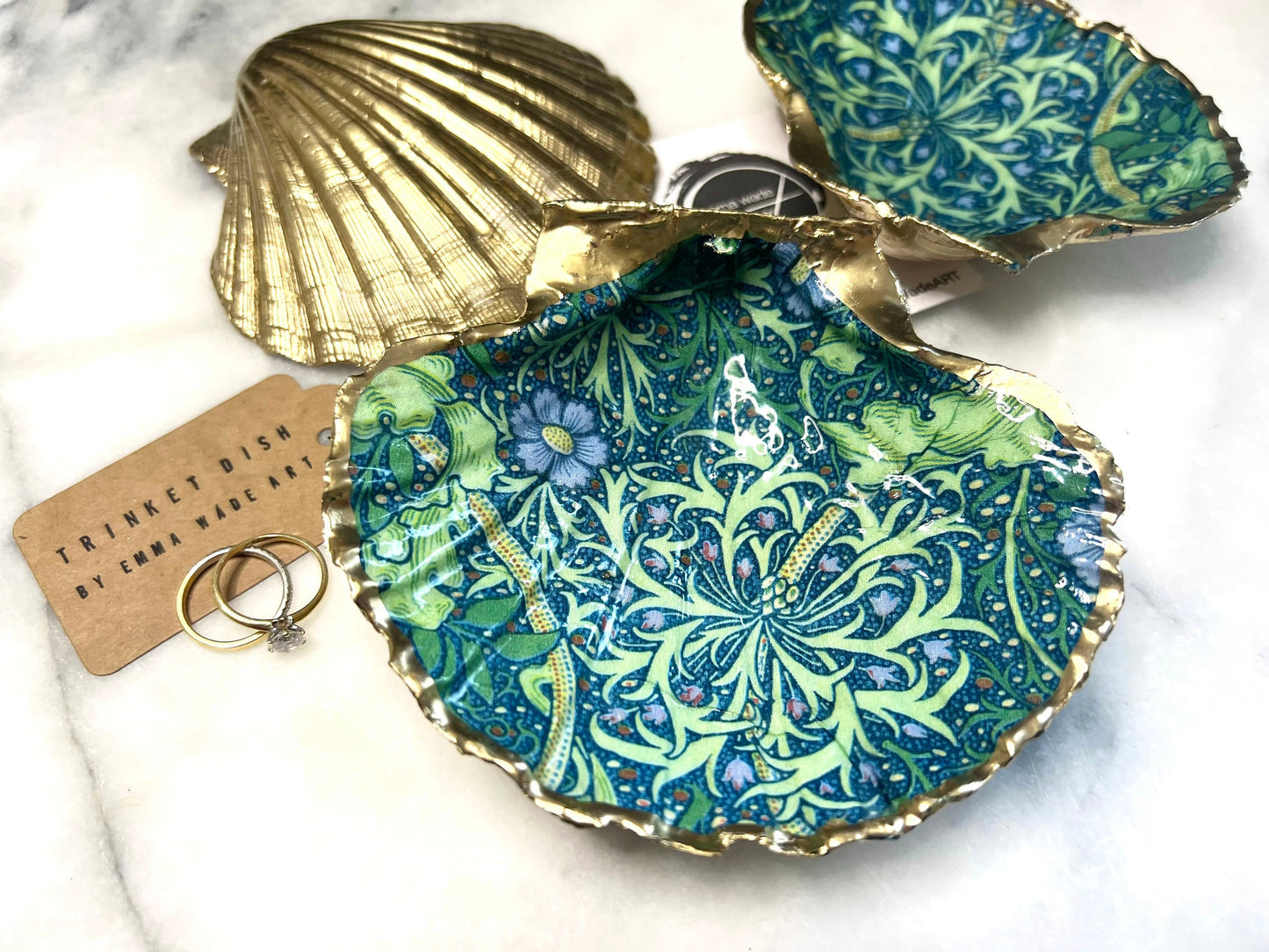 Handmade William Morris inspired scallop shell trinket dish in green and blue tones, featuring a seaweed decoupage design. Finished with gold leaf and sealed with glossy resin. Coastal jewellery dish made from a UK scallop shell, perfect for holding rings, earrings and small treasures. Decorative shell bowl photographed on a light background, showing the natural shell shape and gilded edges.